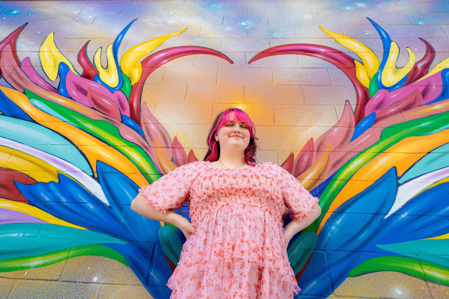 Smiling woman with pink hair in a floral dress posing in front of a colorful heart-shaped mural.