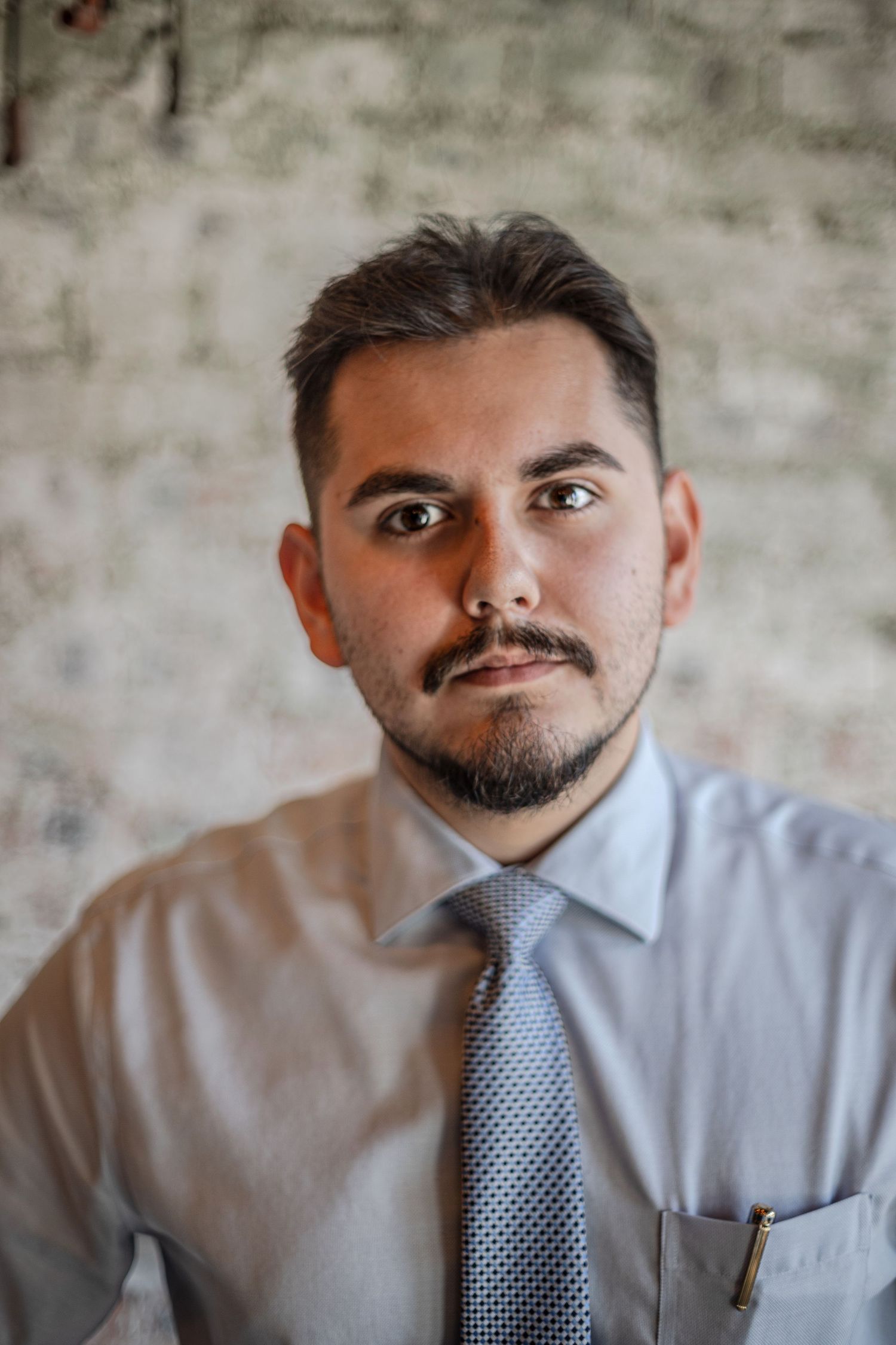 Young bearded man in white dress shirt and tie posing against brick wall.