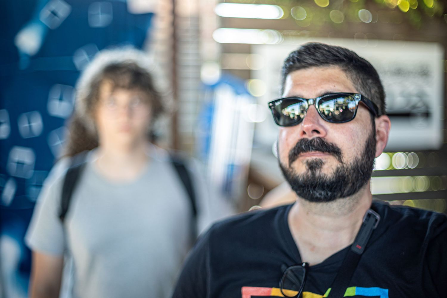 Bearded man wearing reflective sunglasses at an outdoor event with a blurred person in the background.