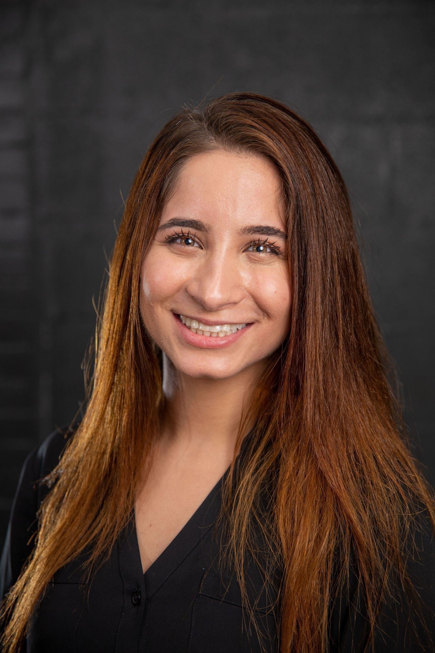Smiling young woman with long auburn hair wearing black scrubs against a dark brick background.