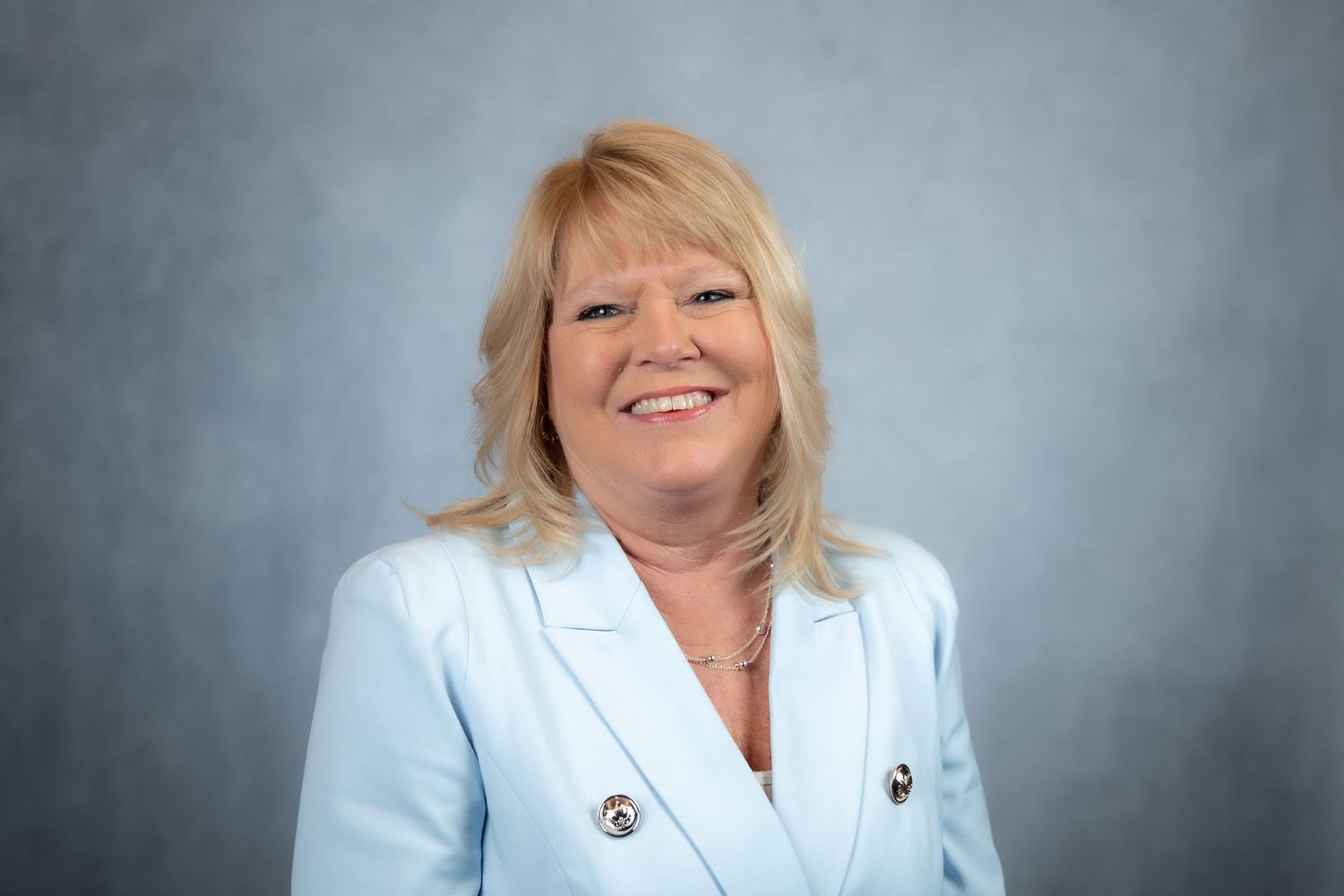 Smiling blonde woman in a light blue blazer posing against a soft gray studio background.