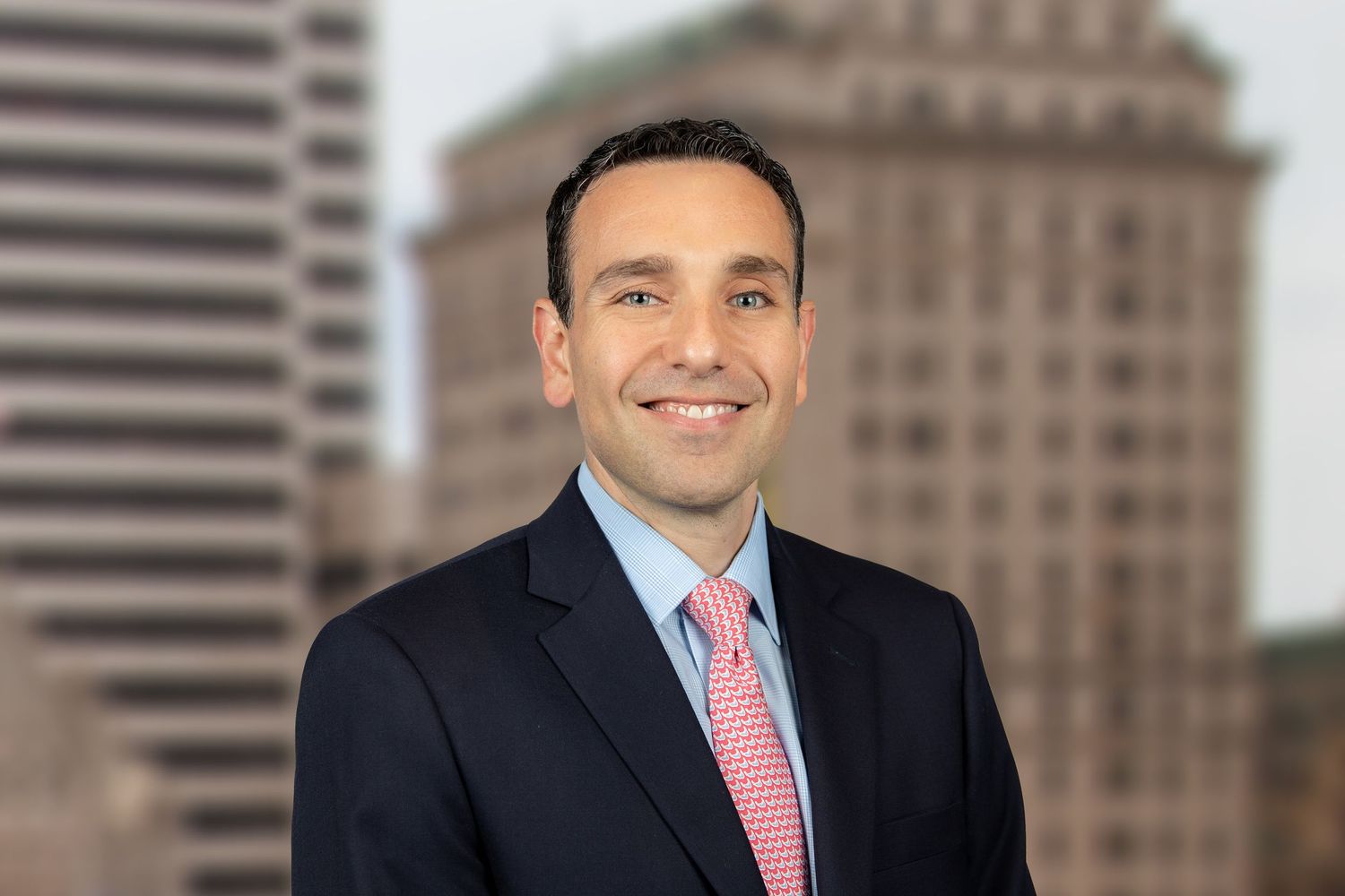 Professional man in dark suit and pink tie smiling with city skyline in background.