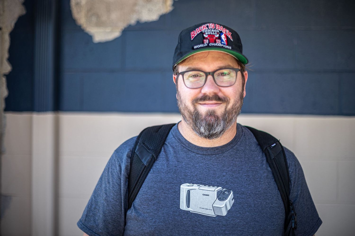 Bearded man wearing glasses and baseball cap smiling outdoors with backpack.