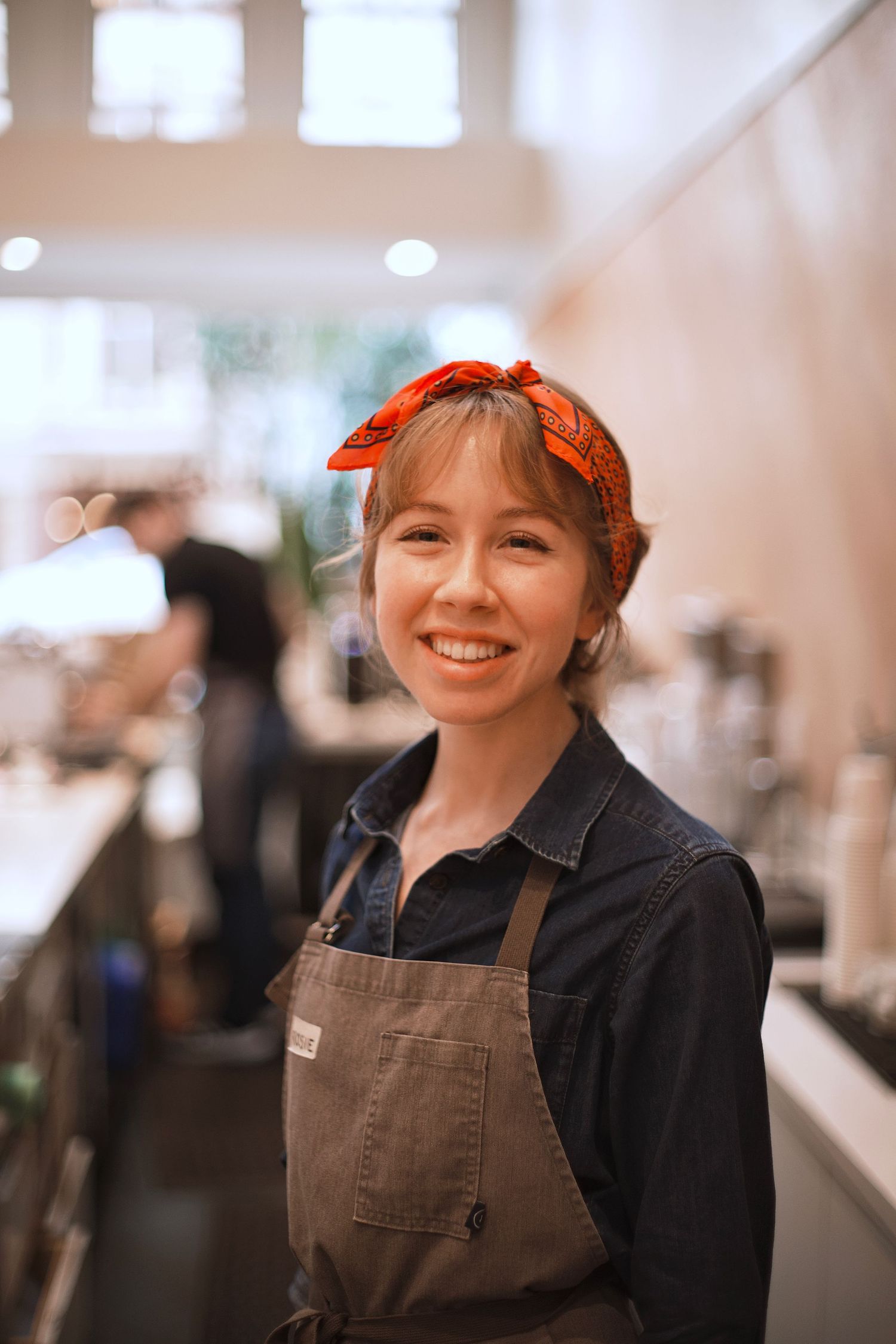 Smiling young woman barista wearing an orange headband and brown apron in a cafe.