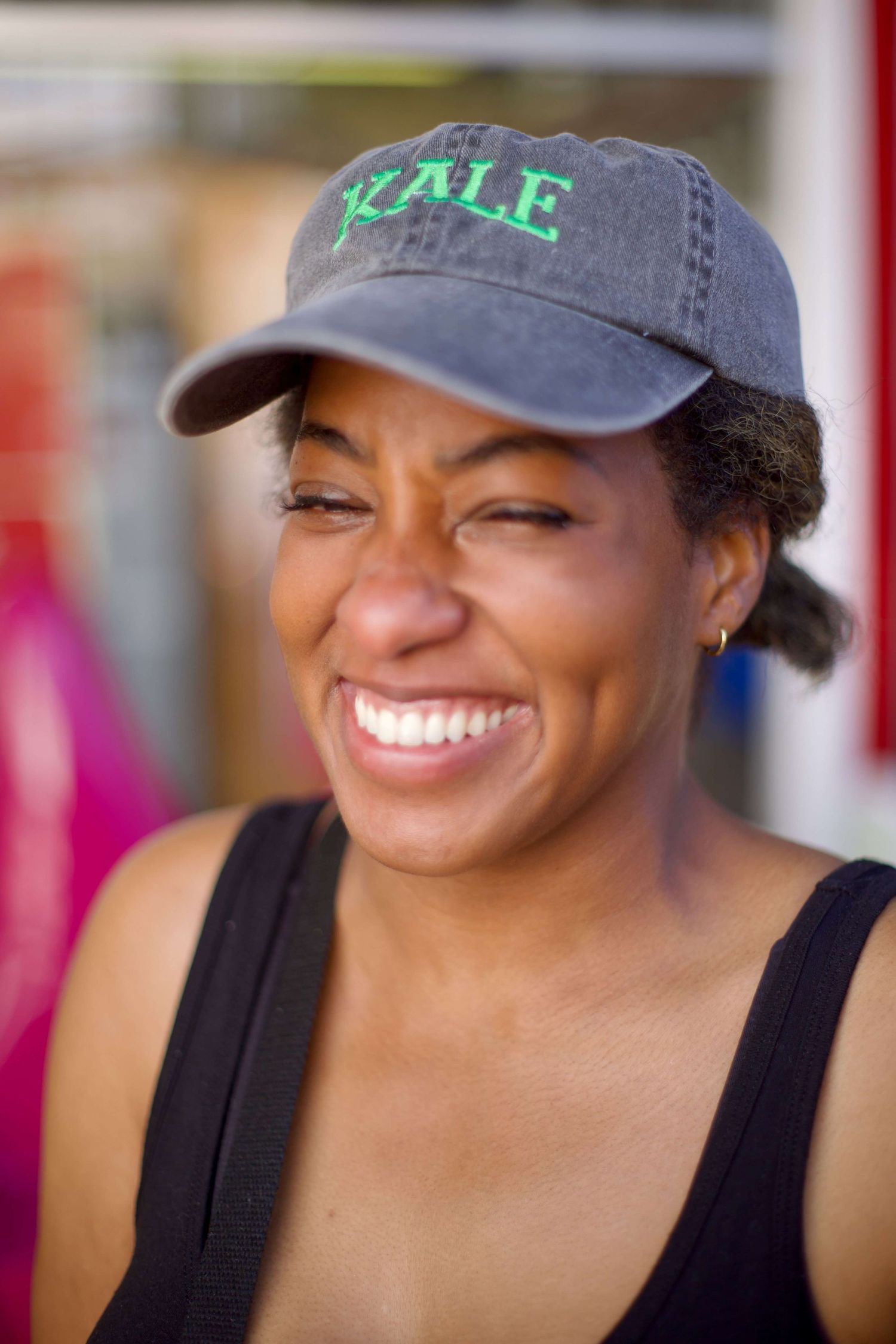 Smiling Black woman wearing a gray KALE baseball cap and black tank top outdoors.