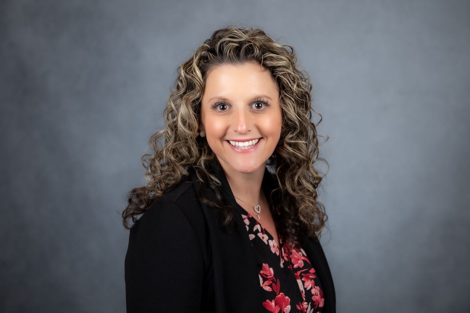 Smiling woman with curly highlighted hair in black blazer poses for professional headshot.