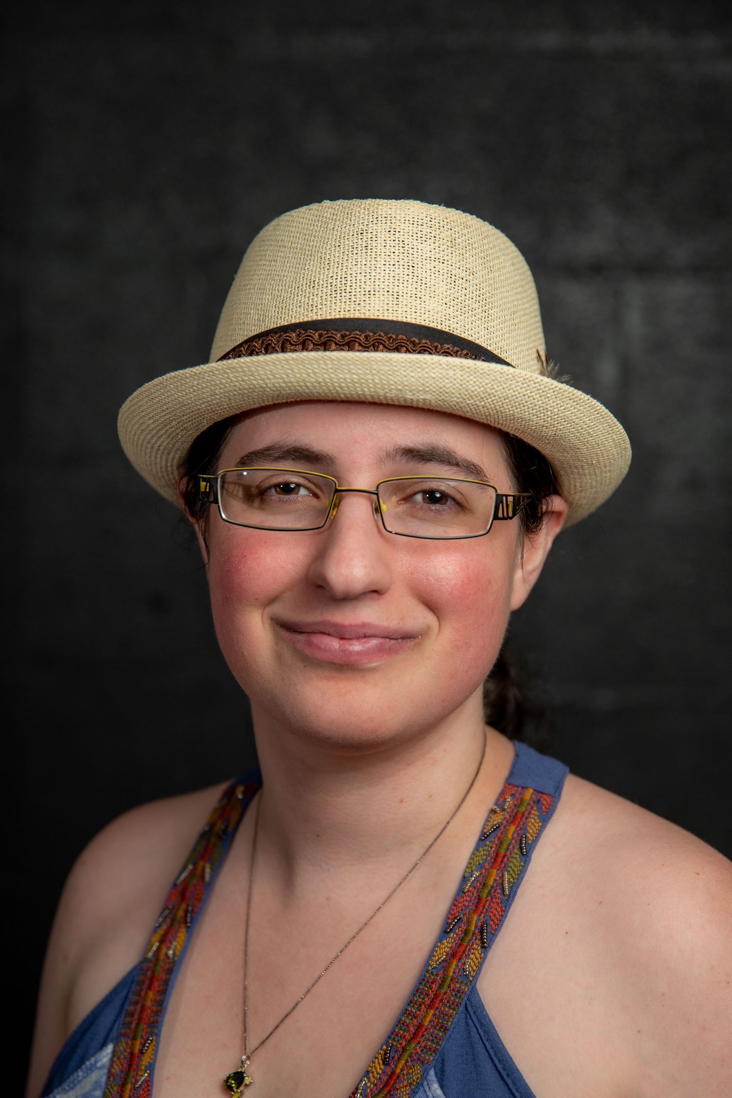 Young woman smiling in a straw fedora hat, glasses, and blue floral top against a dark background.