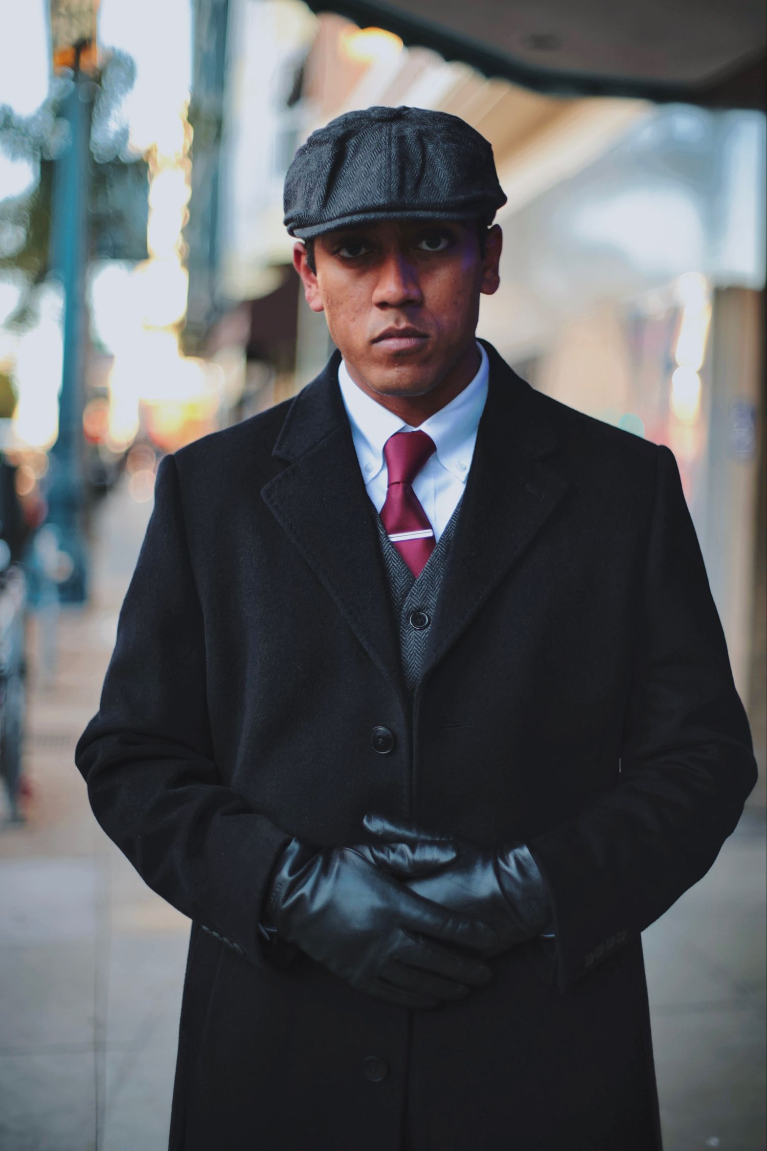 Stylish man in black overcoat, newsboy cap, red tie, and leather gloves poses on city street.