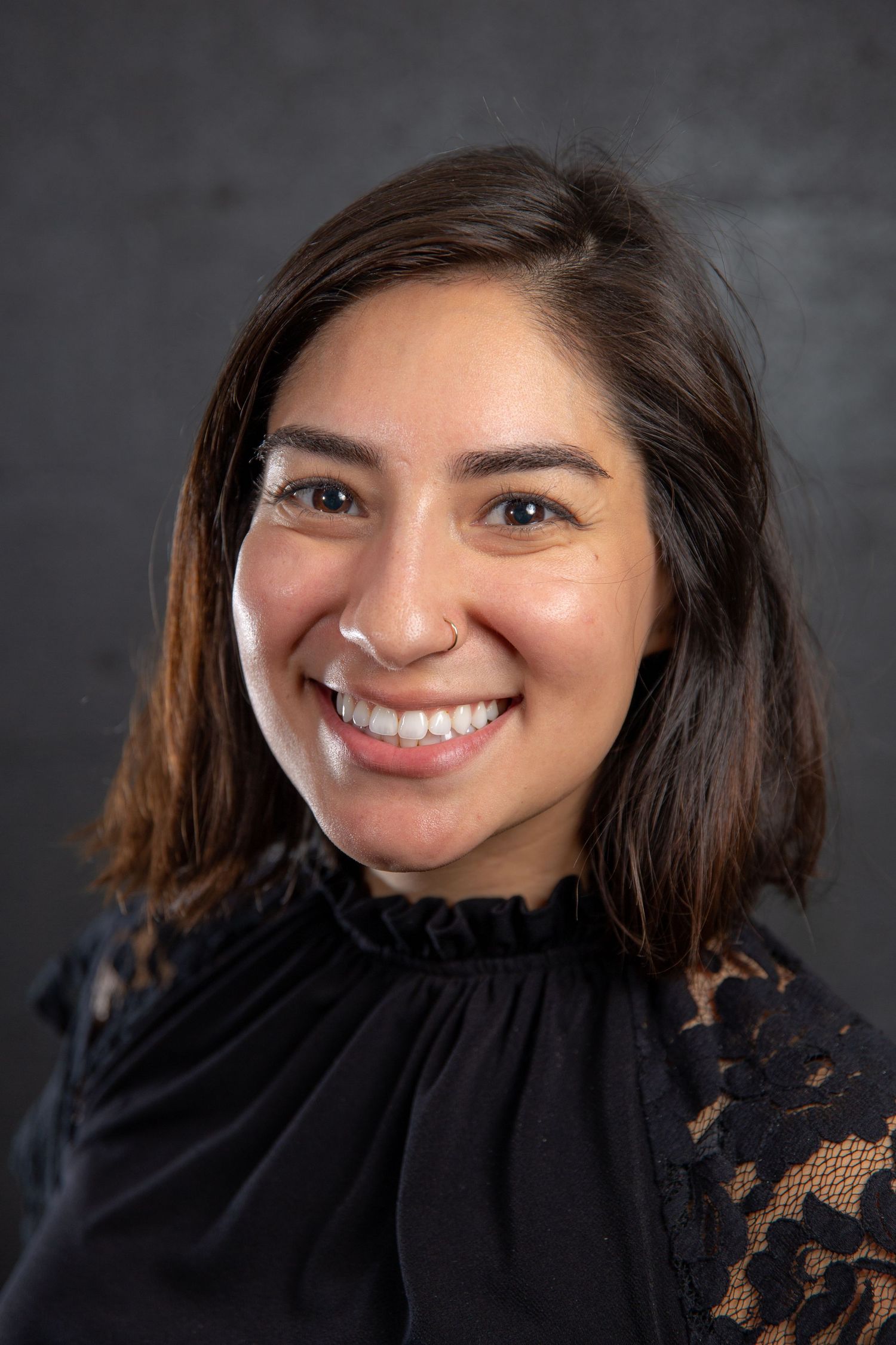 Smiling young woman with brown hair wearing a black lace top, professional headshot on dark background.
