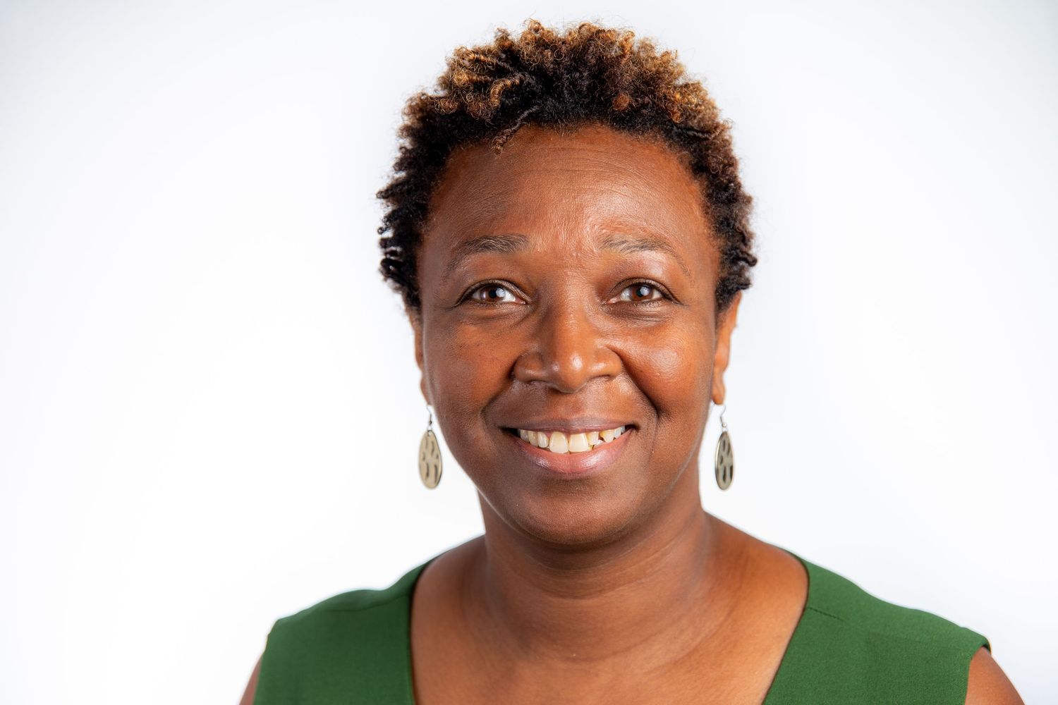 Smiling woman with short natural hair wearing green top and gold earrings on white background.