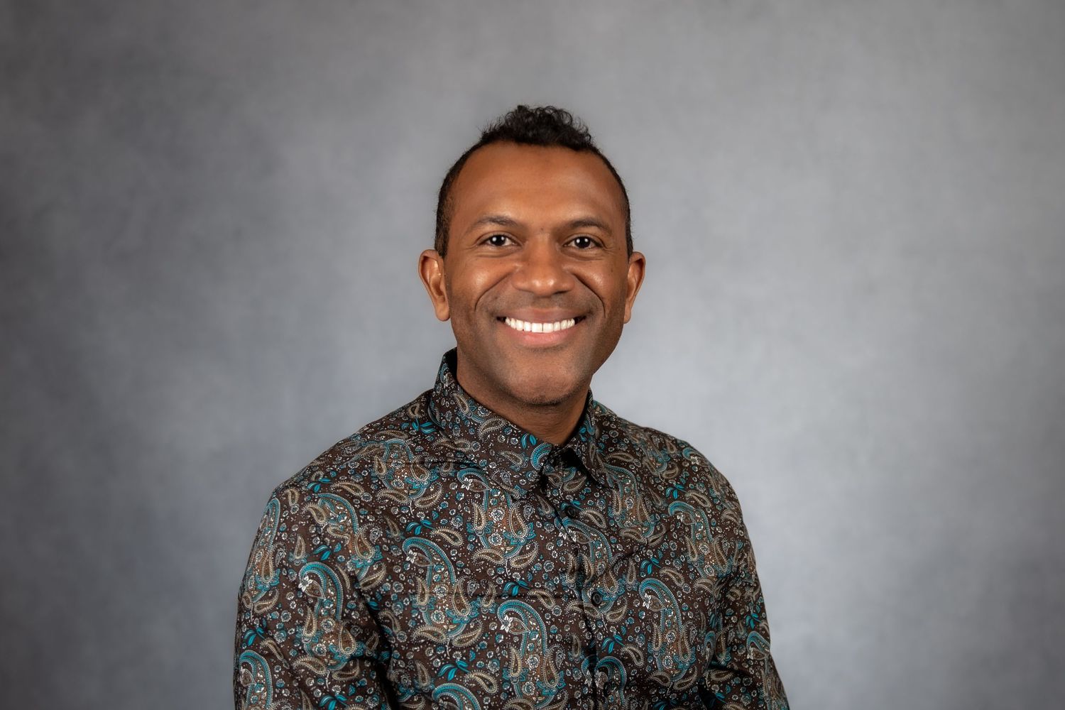 Smiling man in a colorful patterned shirt posing against a gray studio background.