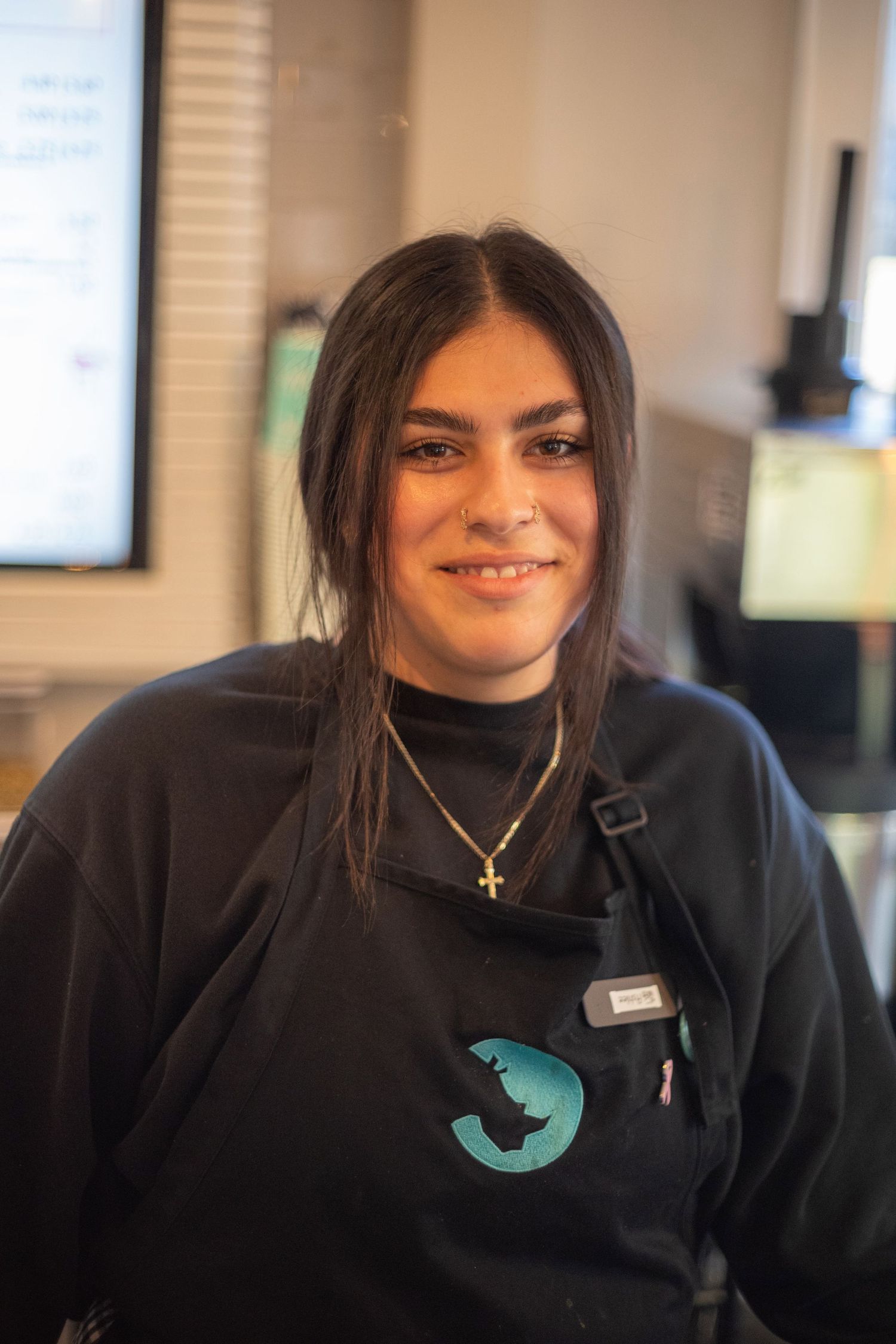 Young woman with dark hair smiling in a black uniform with a cross necklace at a cafe counter.