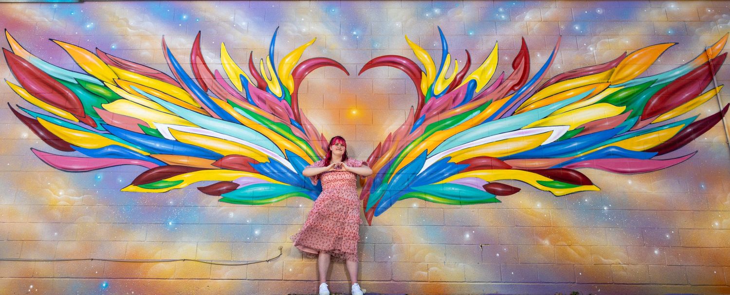 Woman posing in front of a colorful rainbow angel wings mural with a heart shape at the center.