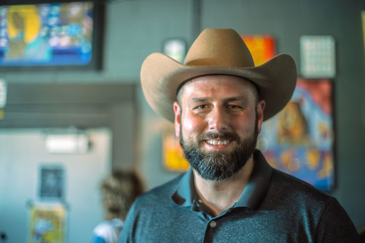 Bearded man wearing a tan cowboy hat and blue polo shirt smiling indoors.
