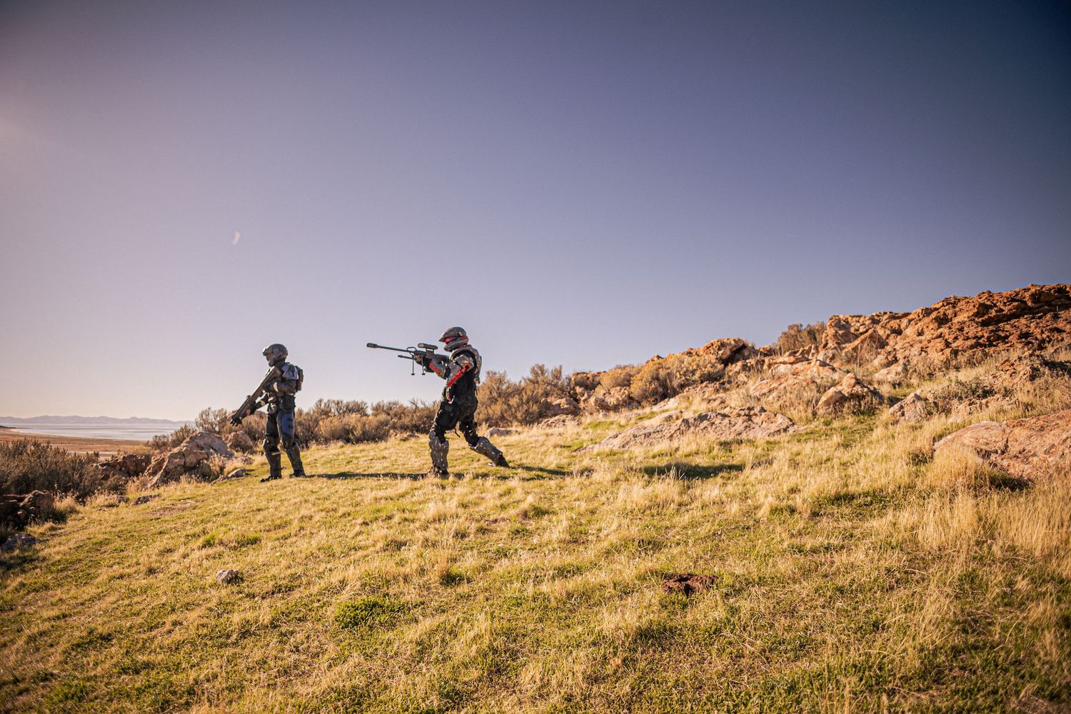 Two Halo cosplayers in black armor practice combat poses on scenic grassy hillside.