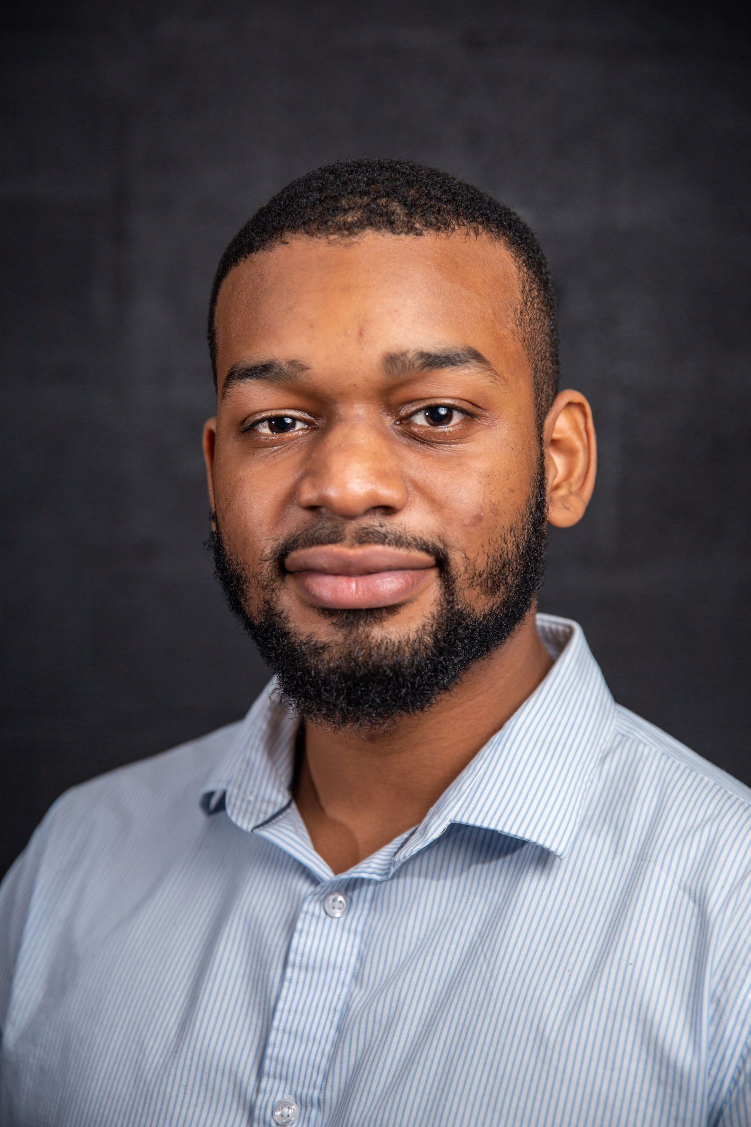 Professional Black man with beard wearing blue striped shirt against dark background.