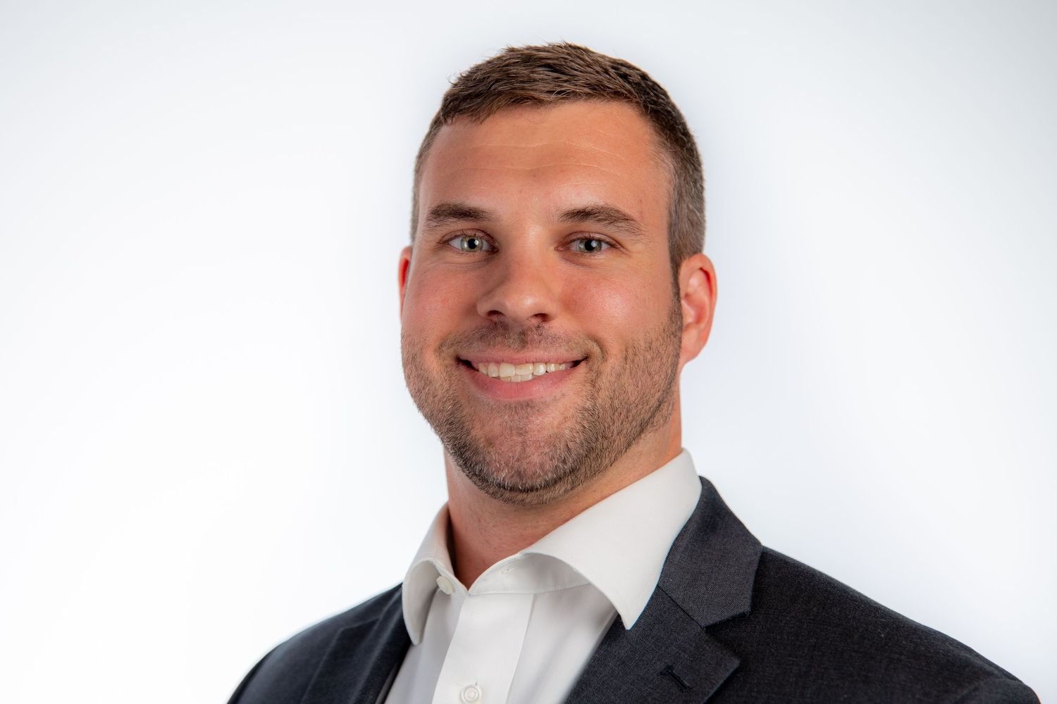 Professional headshot of smiling young man in gray blazer and white shirt on white background.