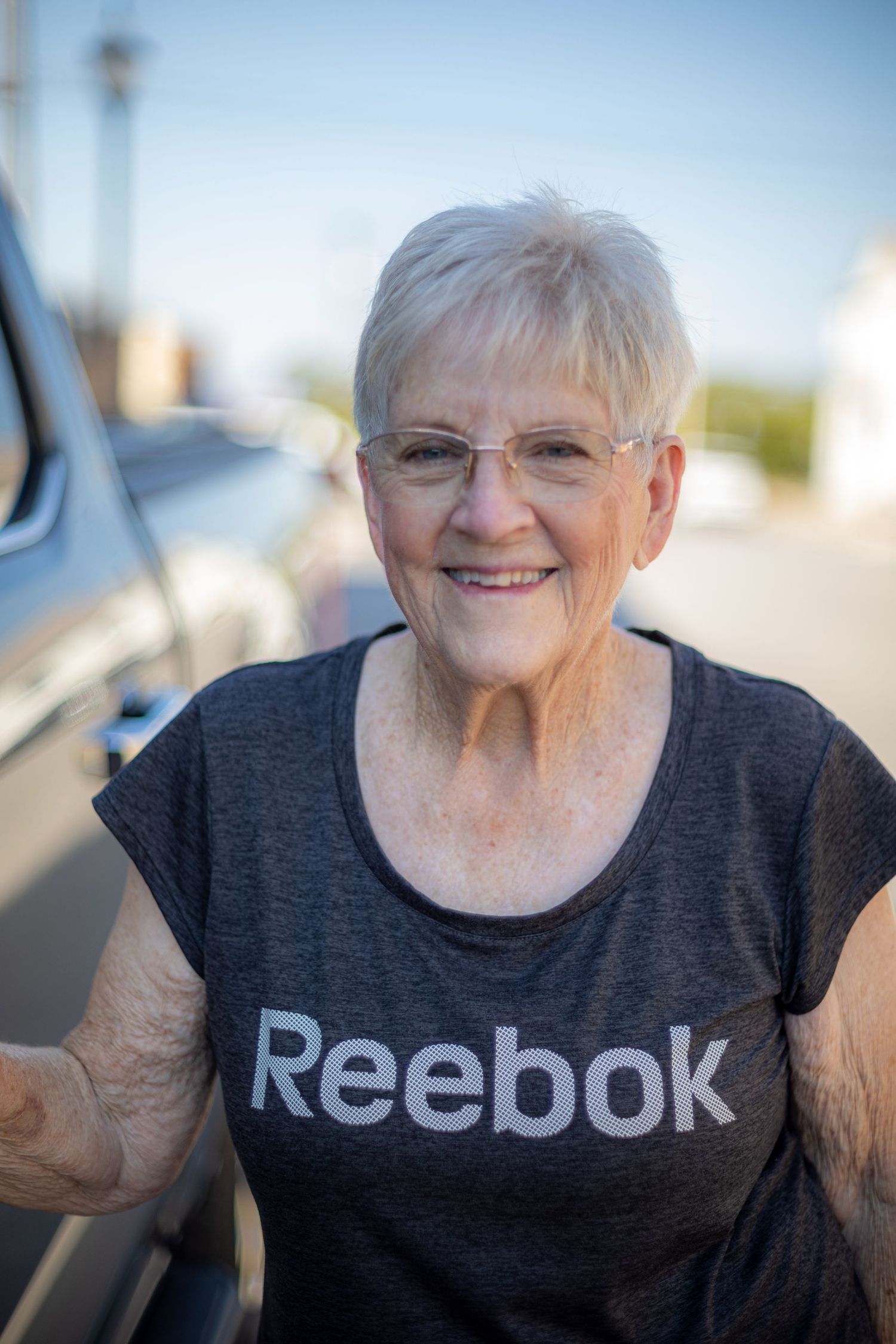 Smiling senior woman with short white hair wearing a gray Reebok t-shirt outdoors near a vehicle.
