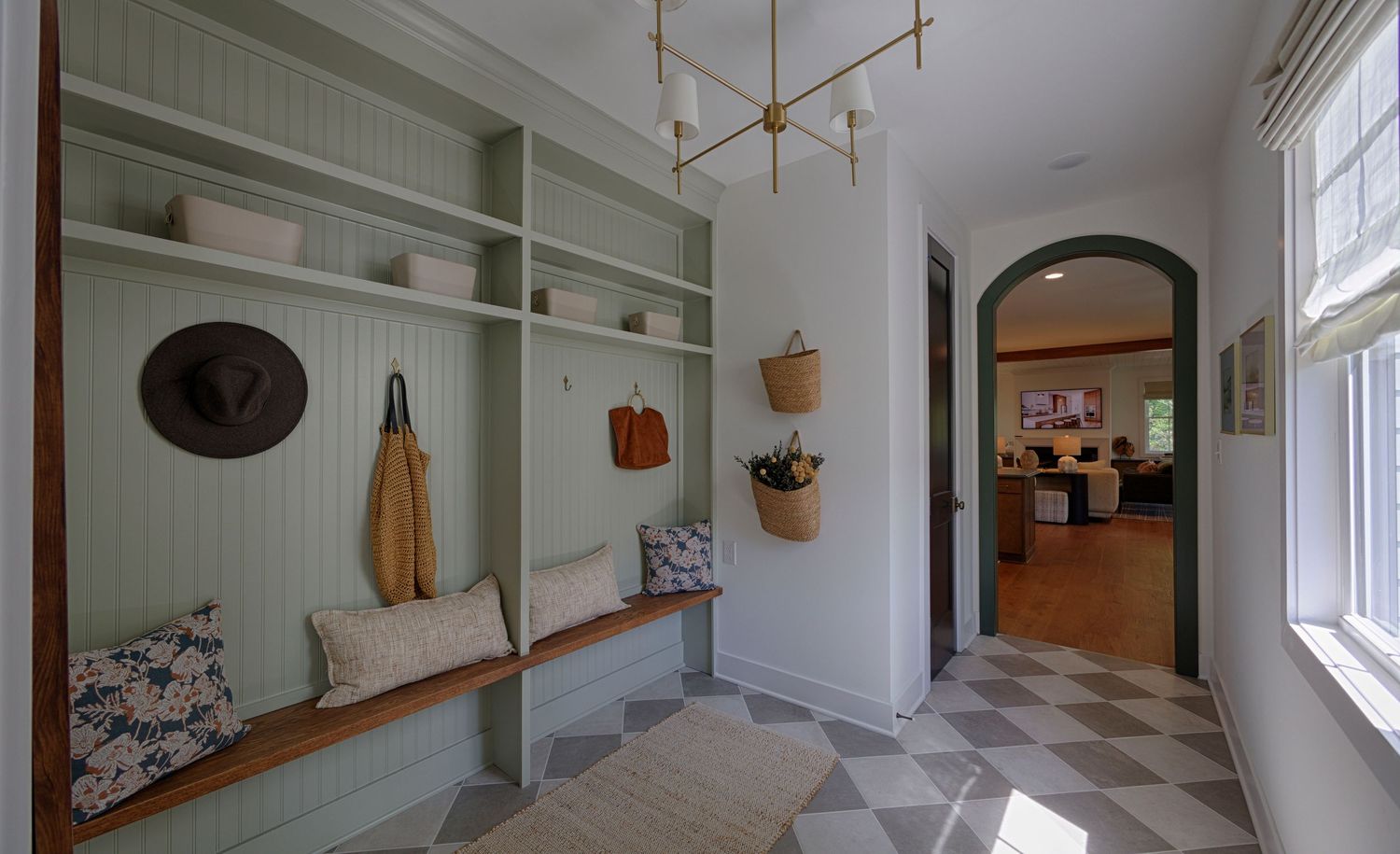 Sage green mudroom with built-in bench, beadboard shelving, woven baskets, checkered floor, and arched hallway leading to living area.