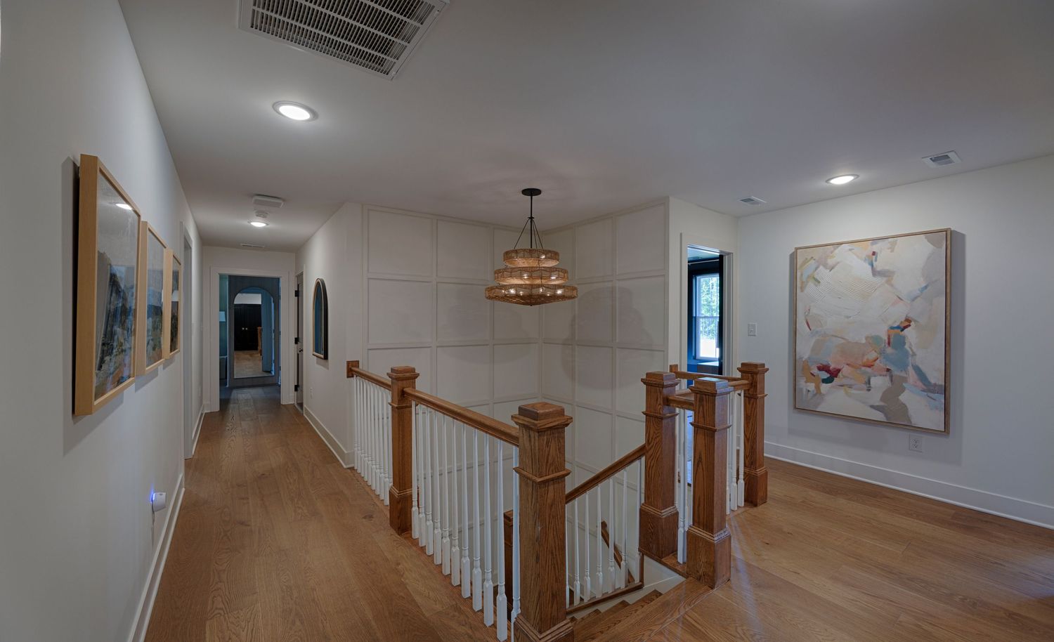 Spacious upstairs hallway with hardwood floors, railing, pendant light, and large abstract art on white walls.