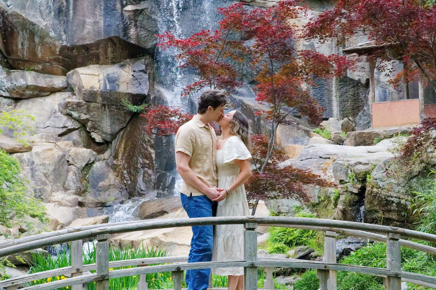 Couple sharing a romantic moment on a bridge near a waterfall surrounded by lush red maple trees.