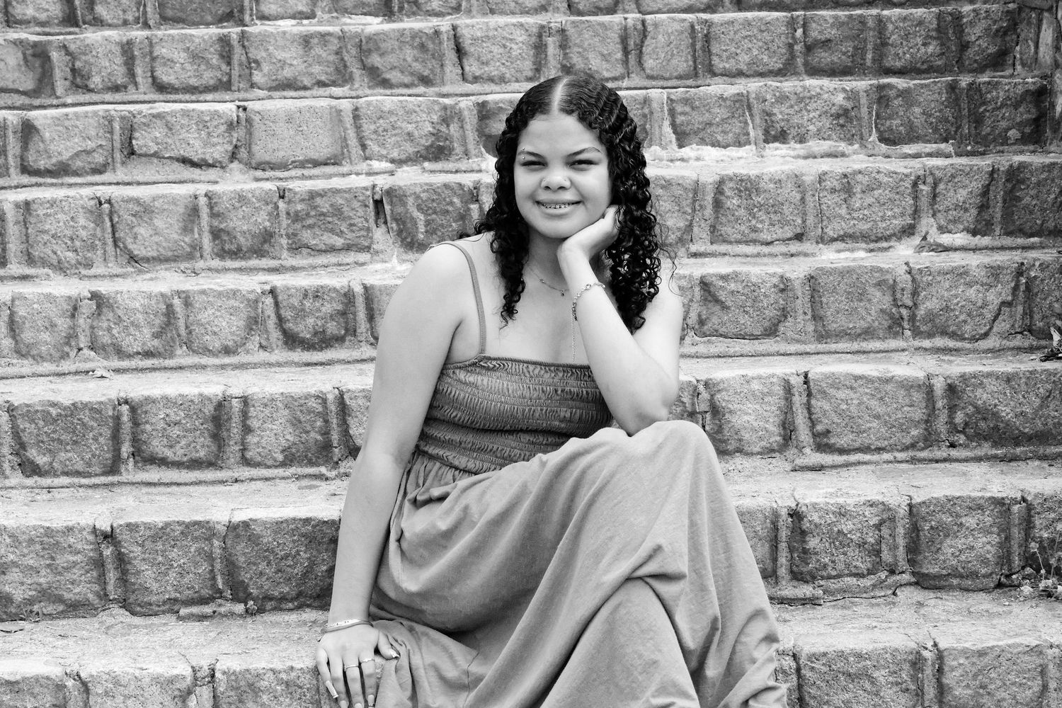 Young woman in elegant gown sitting gracefully on stone steps, posing with hand on chin in black and white photo.