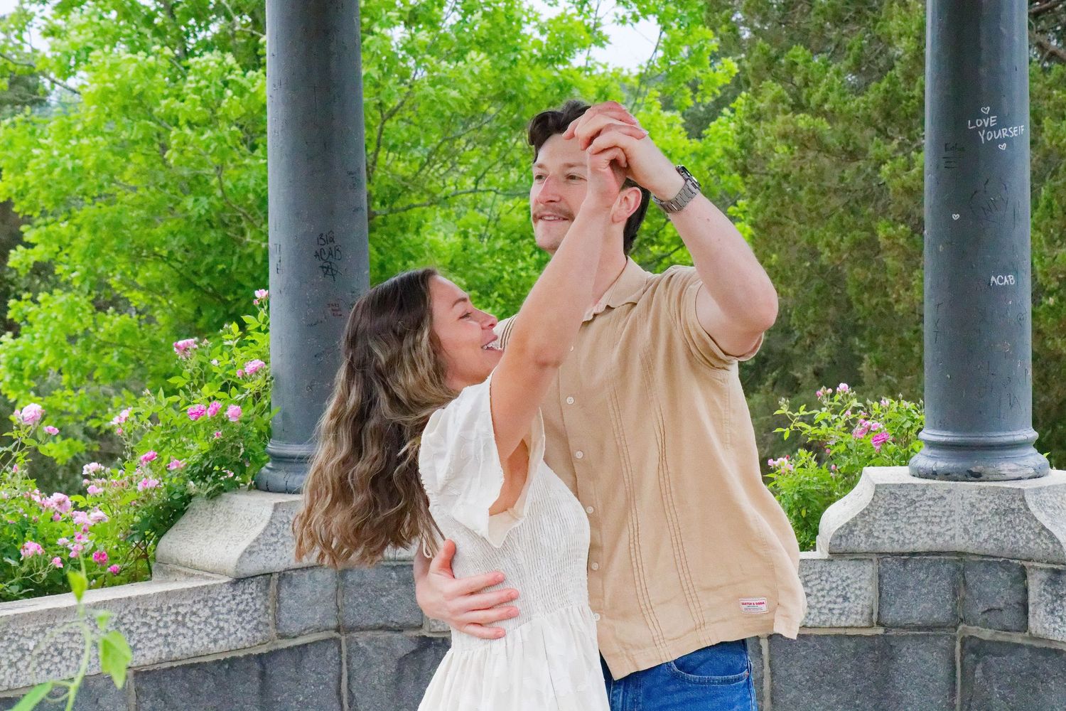 A couple dances together outdoors near stone columns surrounded by lush green trees and blooming flowers.