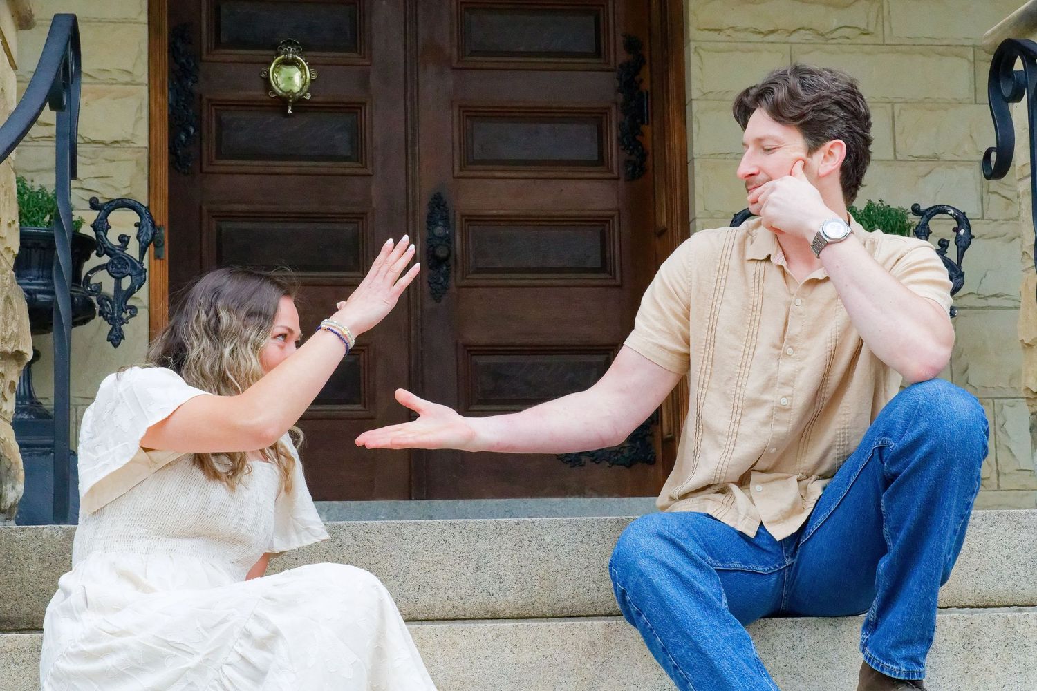 Couple sitting on stone steps laughing and gesturing in front of elegant wooden doors.