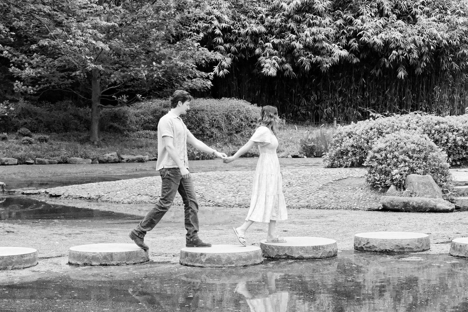 Couple holding hands crossing stepping stones over a pond in a lush garden, black and white photo.