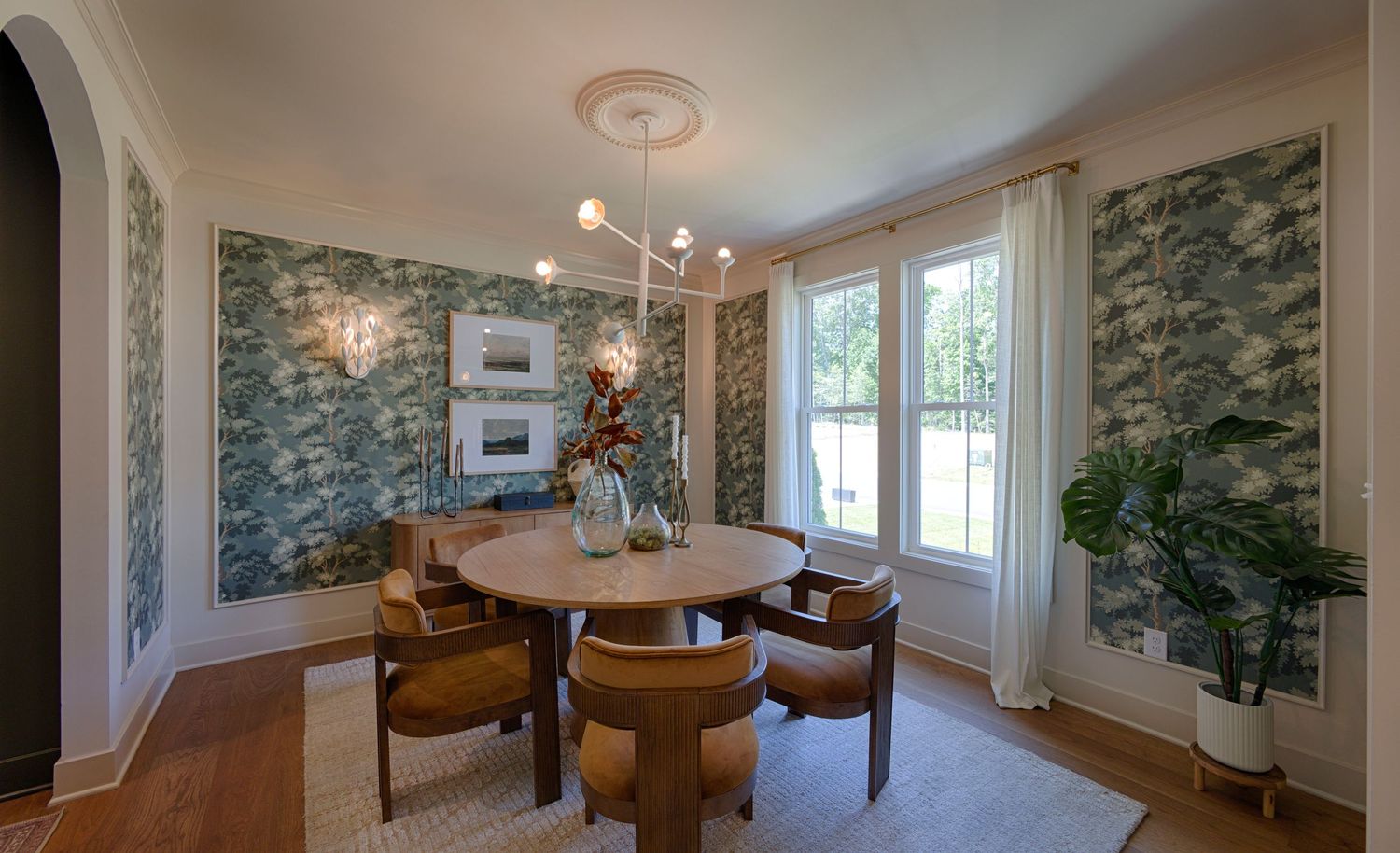 Elegant dining room with botanical wallpaper, round wood table, tan chairs, modern chandelier, and large windows with natural light.