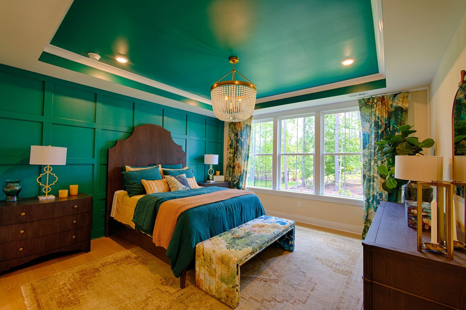 Luxurious teal bedroom with tray ceiling, chandelier, upholstered bed, and large window with garden views.Richmond, Virginia.
