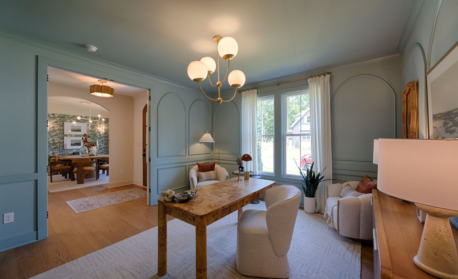 Elegant blue-green dining room with wainscoting, chandelier, wooden table, and arched doorway leading to a cozy living space.