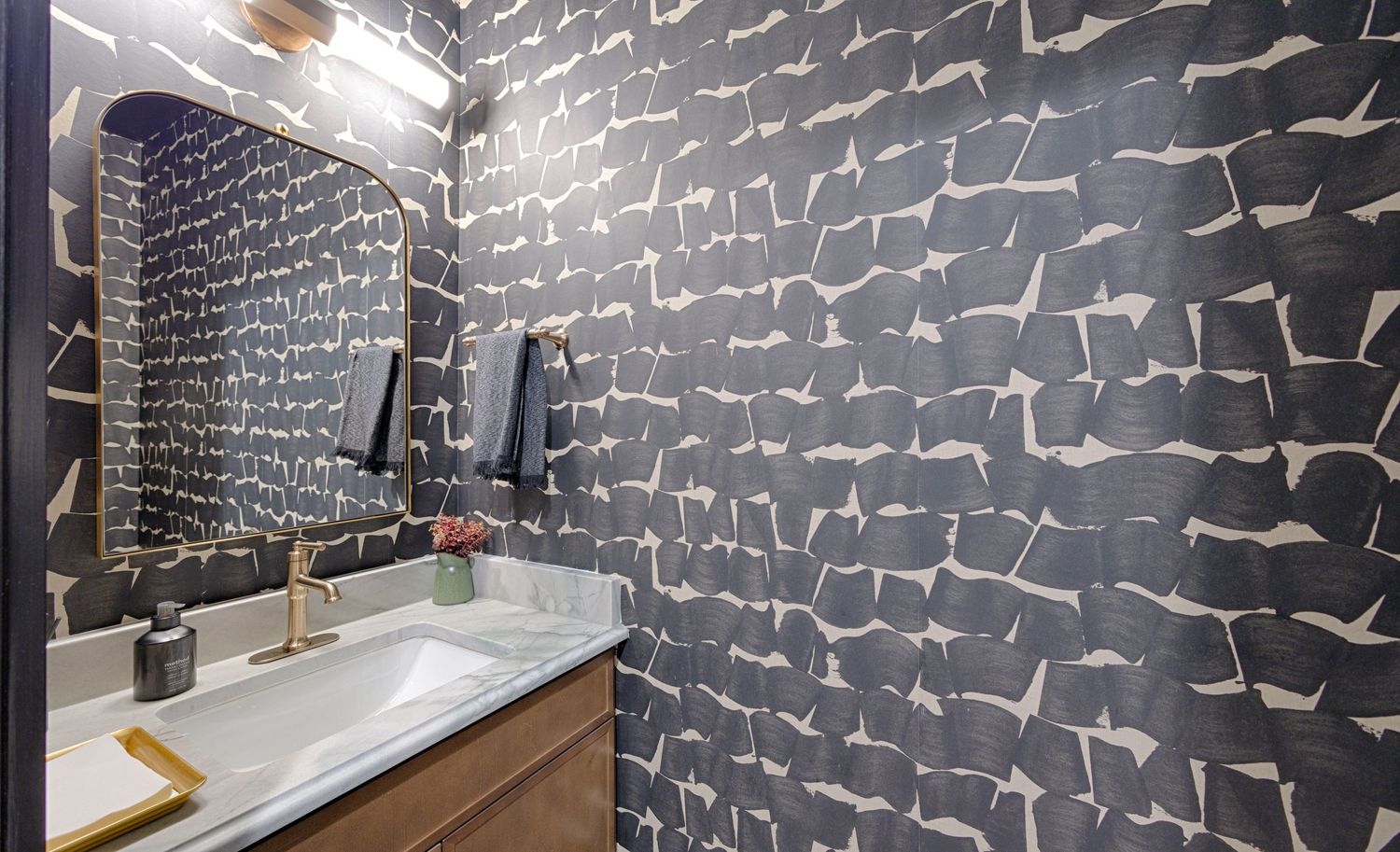 Modern powder room featuring bold dark patterned wallpaper, gold-accented vanity, white sink, and backlit mirror.