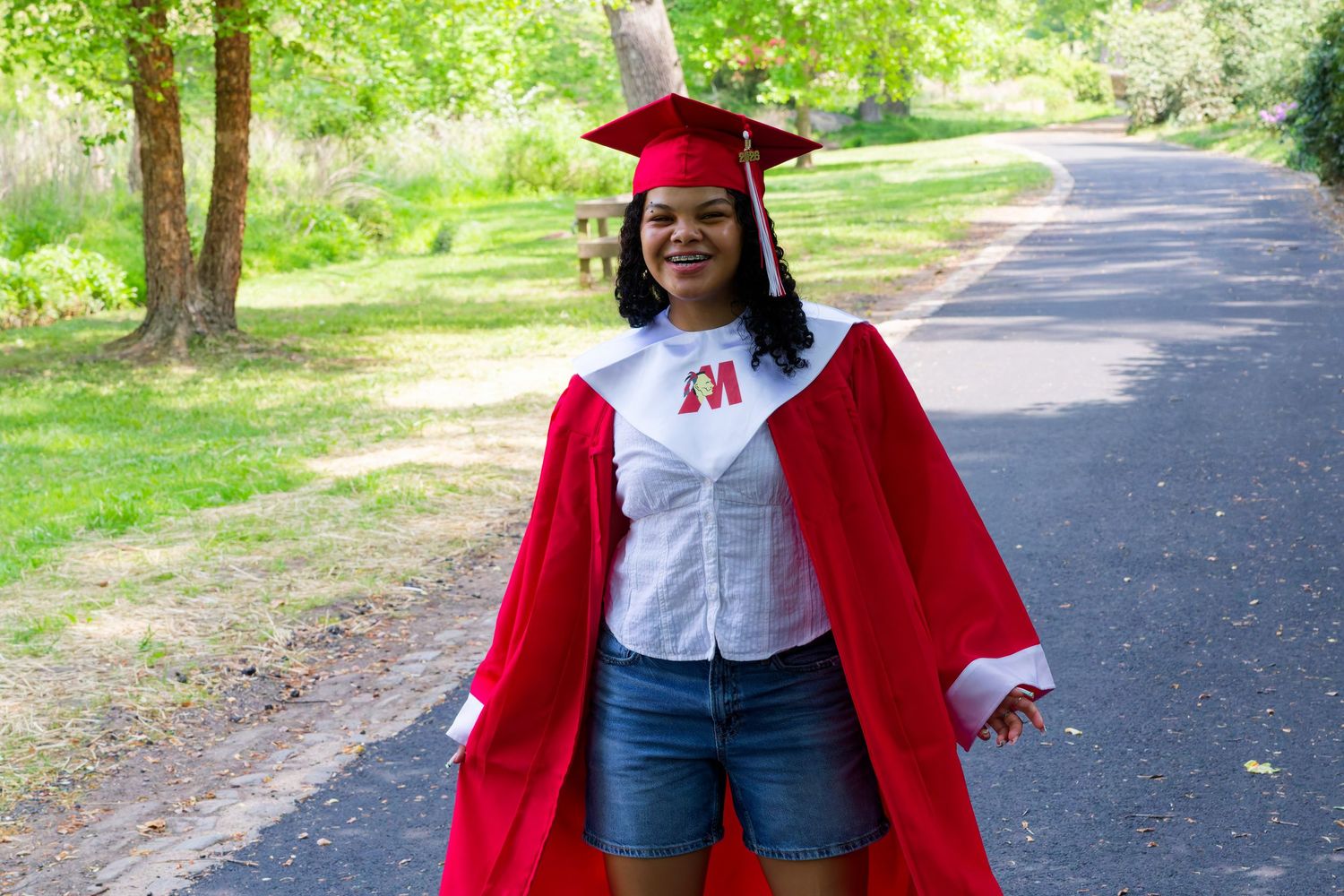 Smiling graduate in red cap and gown stands on a tree-lined path celebrating her graduation day.