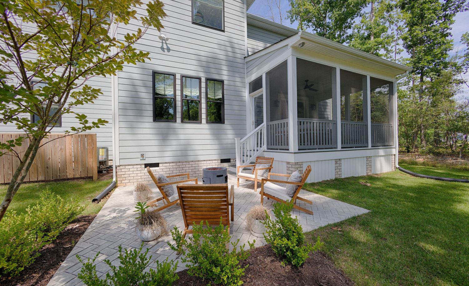 Backyard patio with Adirondack chairs around a fire pit, lush lawn, and screened porch on a white suburban home.