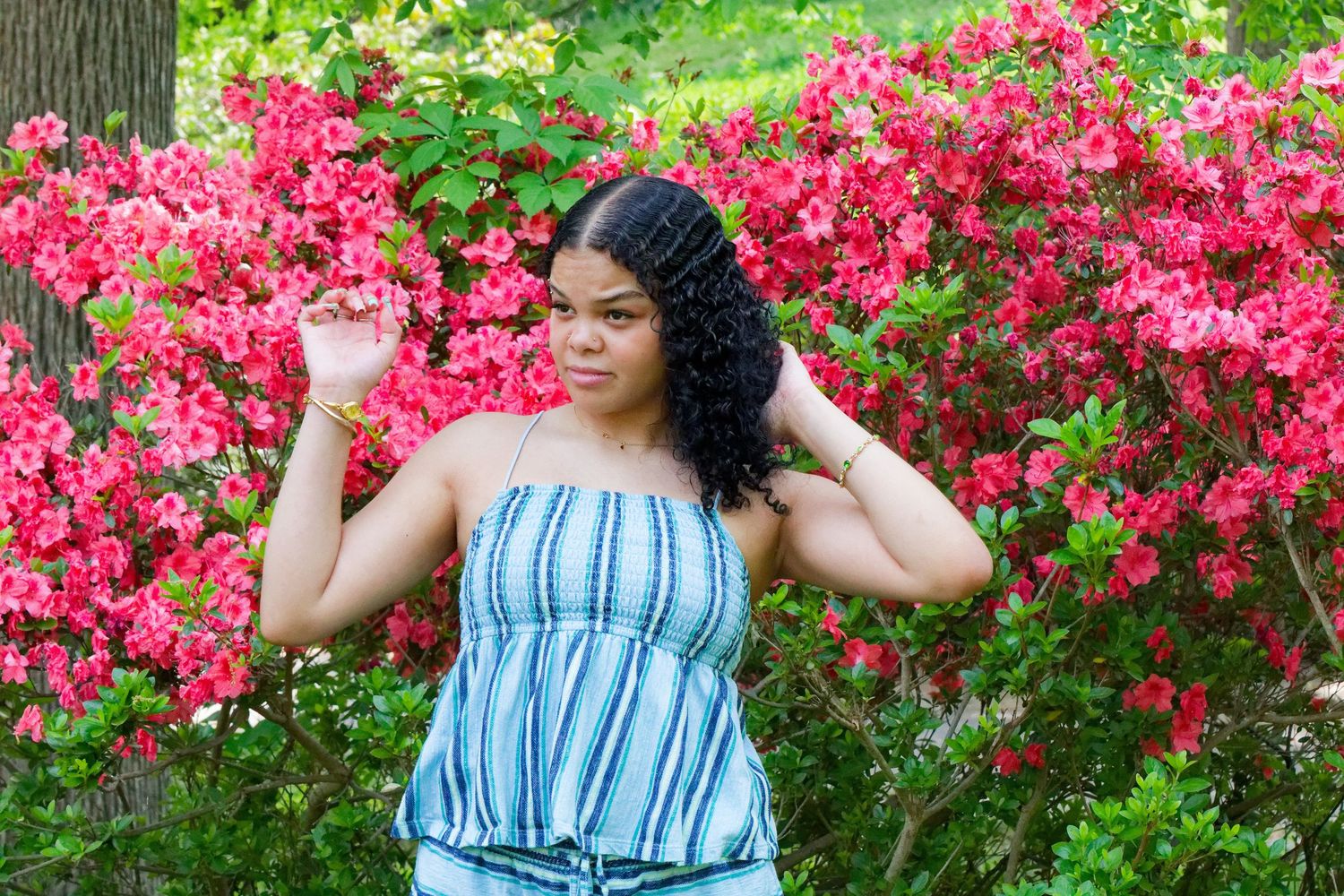 Young woman in striped tube top posing gracefully among vibrant pink azalea bushes in a lush garden.