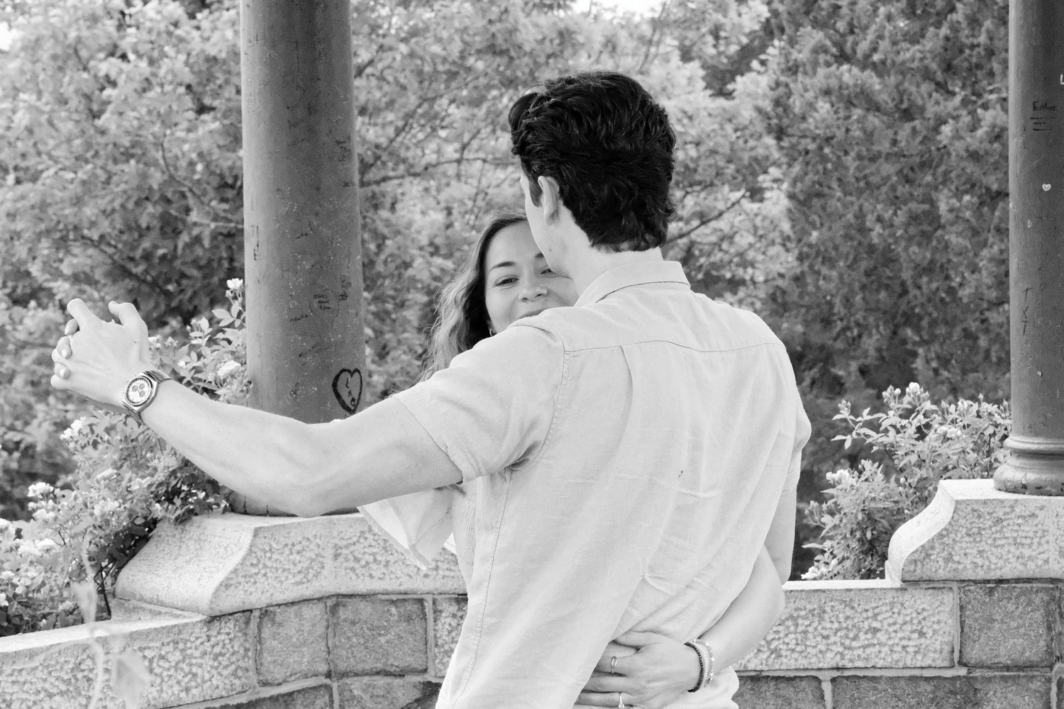 Black and white photo of couple embracing near stone columns surrounded by lush greenery.