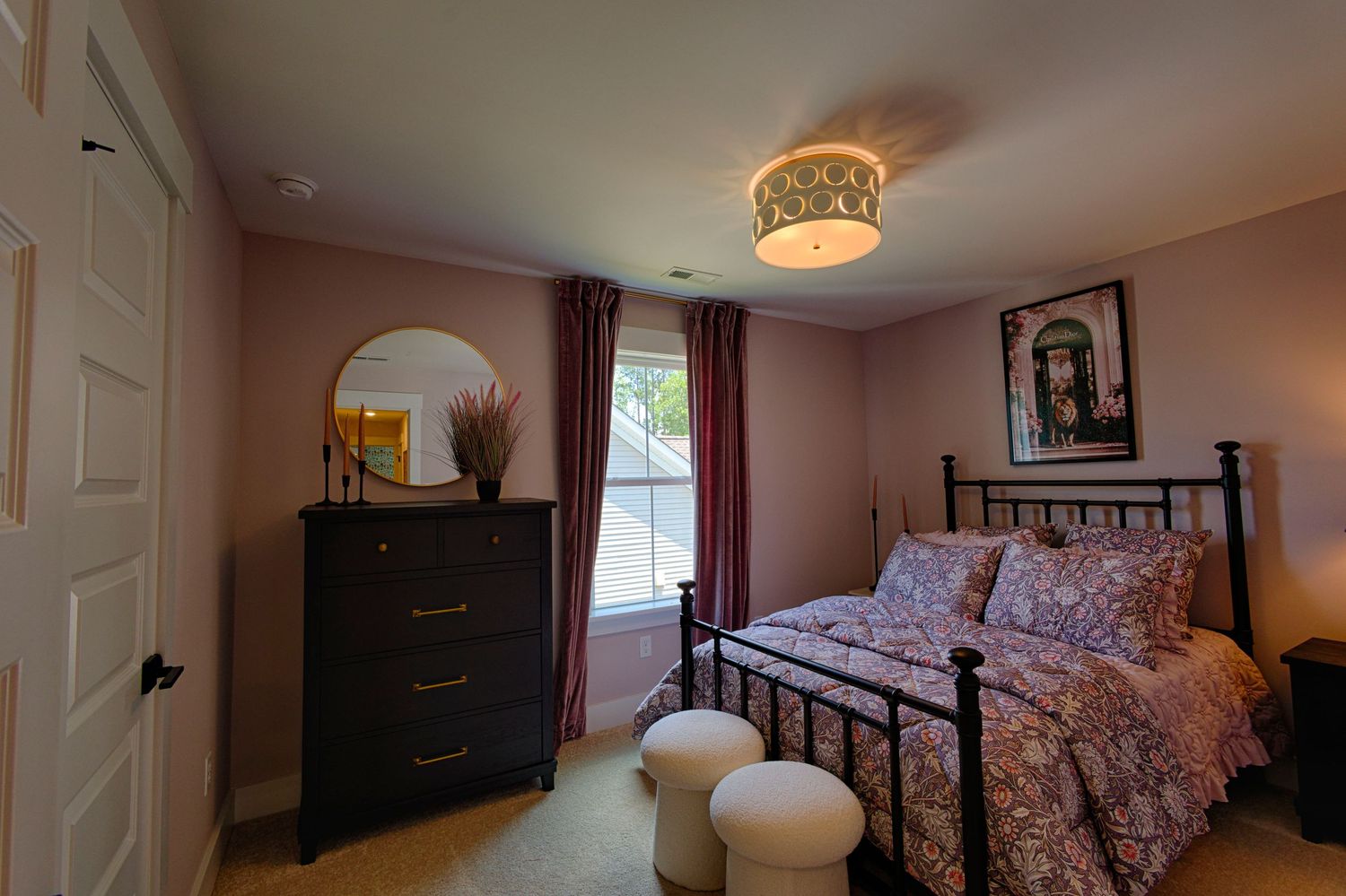 Cozy bedroom with iron bed frame, floral bedding, dark wood dresser with mirror, and soft pink walls with warm lighting.Richmond, Virginia.