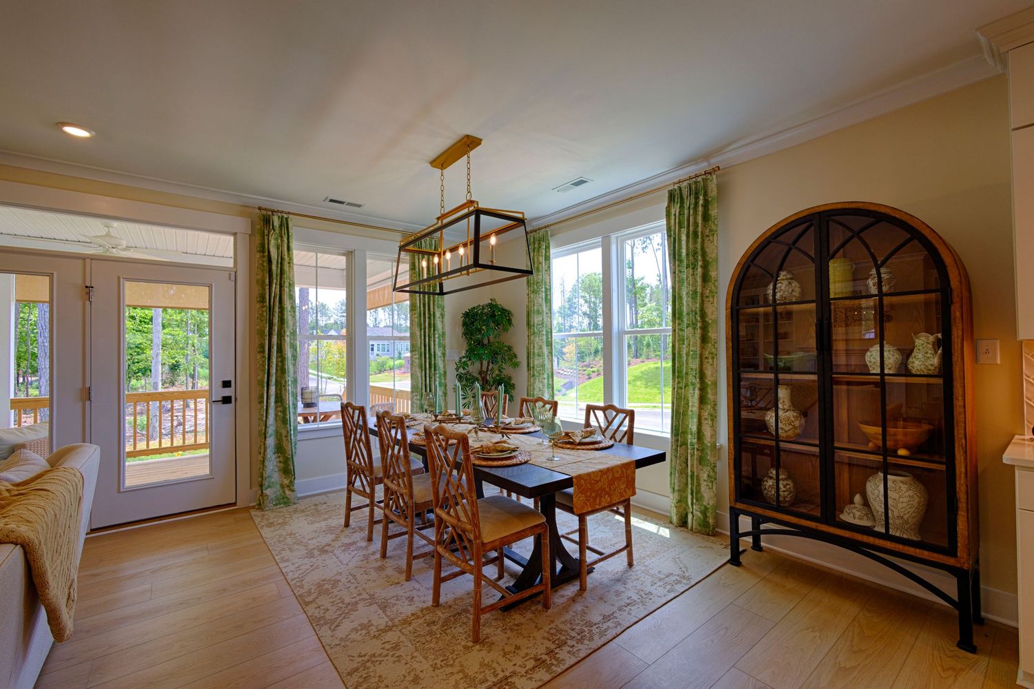 Bright dining room with wooden table, chairs, arched china cabinet, green curtains, and large windows overlooking a lush yard. Real Estate