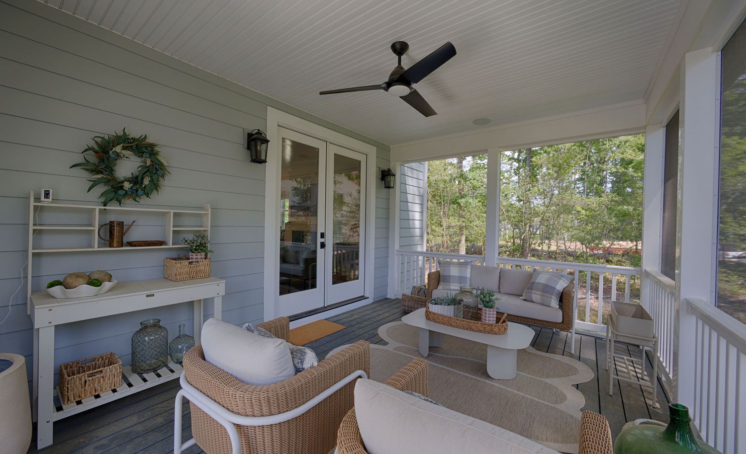 Cozy covered porch with wicker furniture, ceiling fan, French doors, and decorative wreath on gray siding.