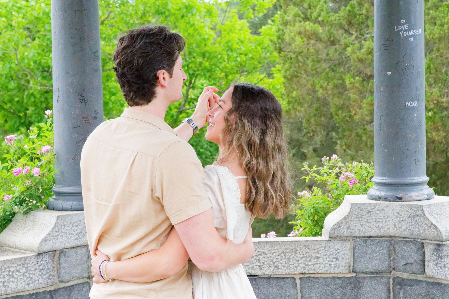 A romantic couple embraces near stone columns surrounded by lush greenery and pink flowers outdoors.