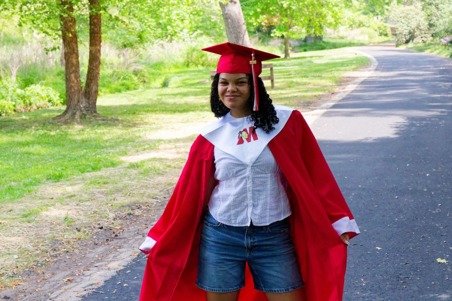 Young woman in red graduation cap and gown smiling on a tree-lined path outdoors.