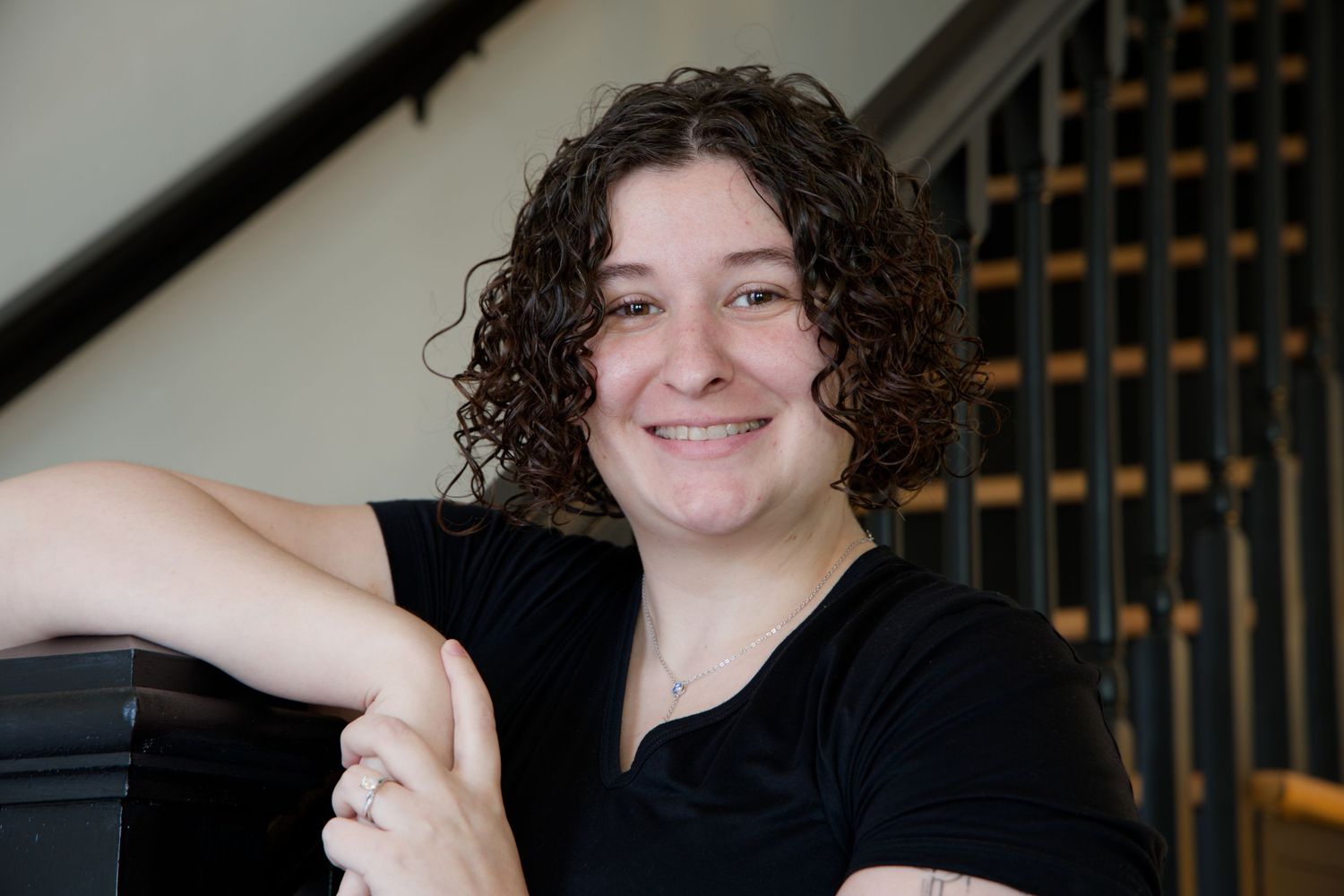 A person with curly dark hair smiles warmly while posing in a black shirt against a light background. Indoor Photography, RVA, Richmond, VA