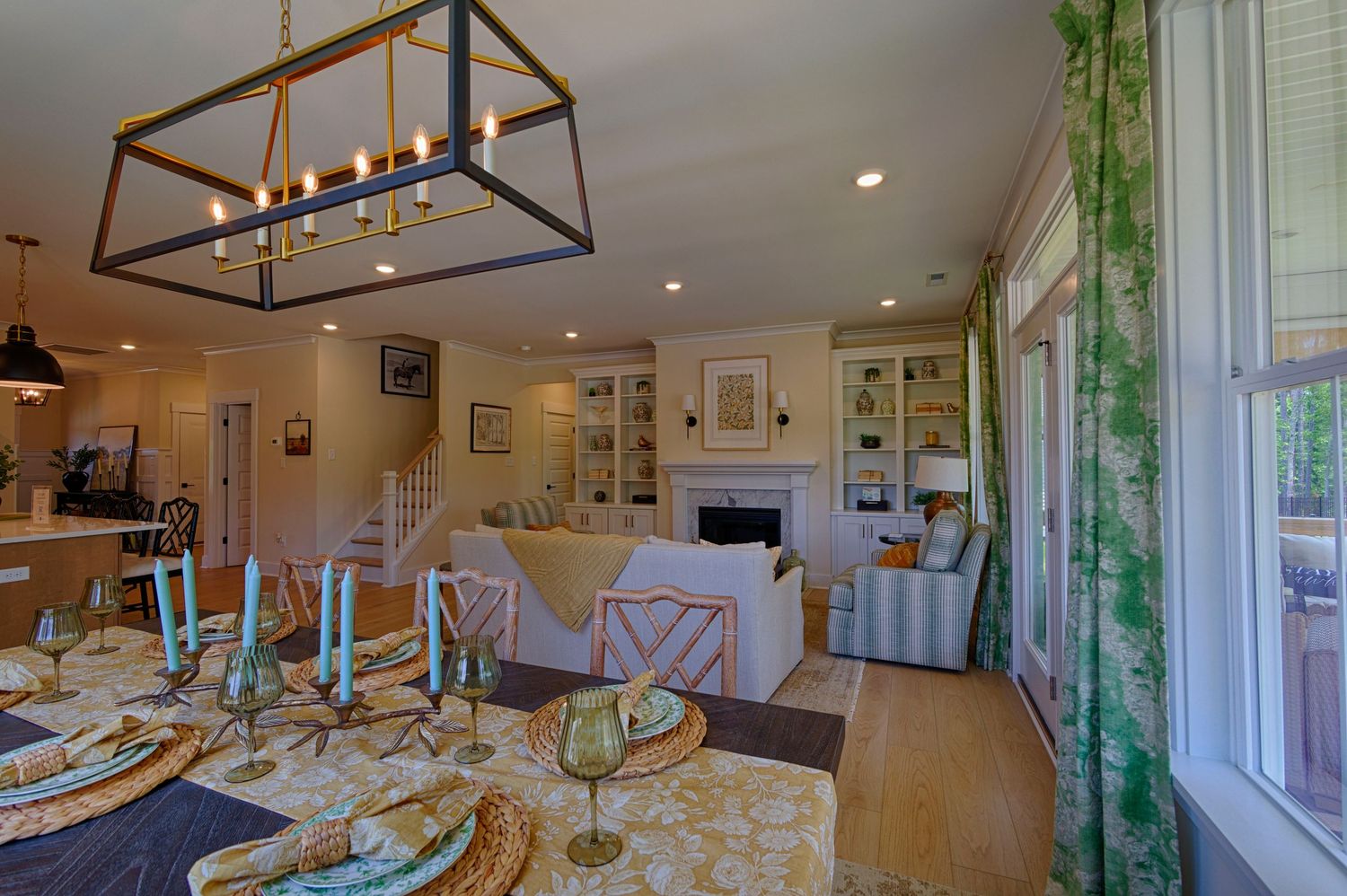 Open-plan dining and living room with modern chandelier, yellow floral accents, white built-ins, and green botanical curtains. Real Estate
