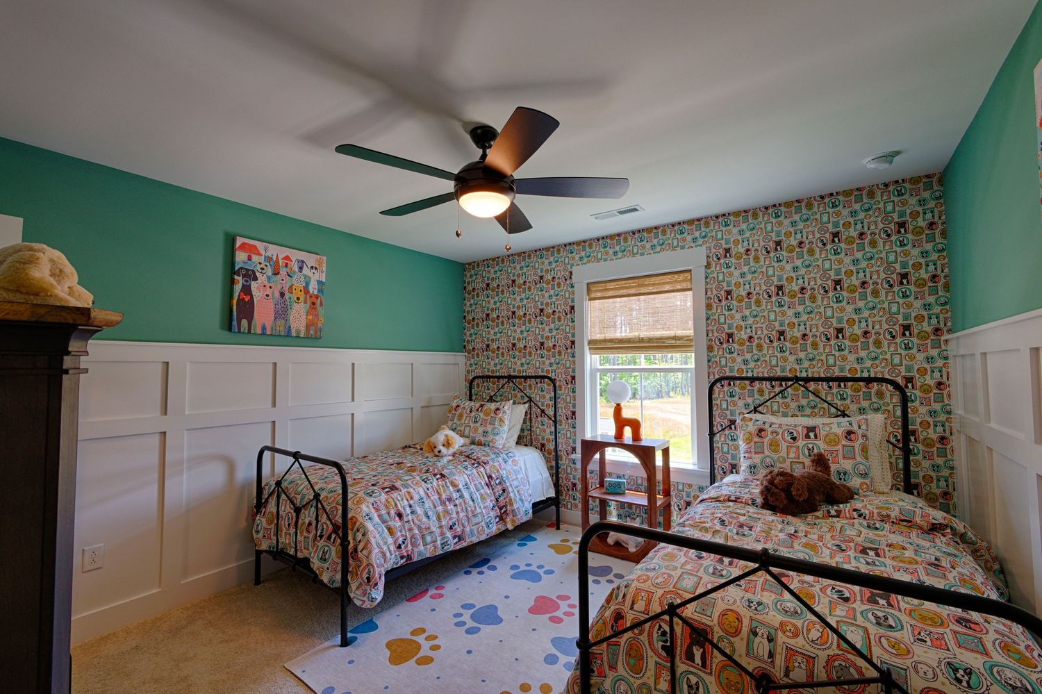 Colorful kids bedroom with two iron beds, teal walls, wainscoting, ceiling fan, and playful patterned wallpaper.Richmond, Virginia.