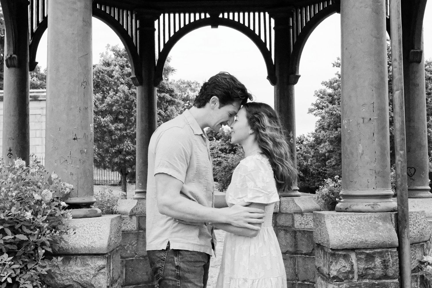 Black and white photo of a couple sharing a romantic moment under a gazebo surrounded by lush greenery.