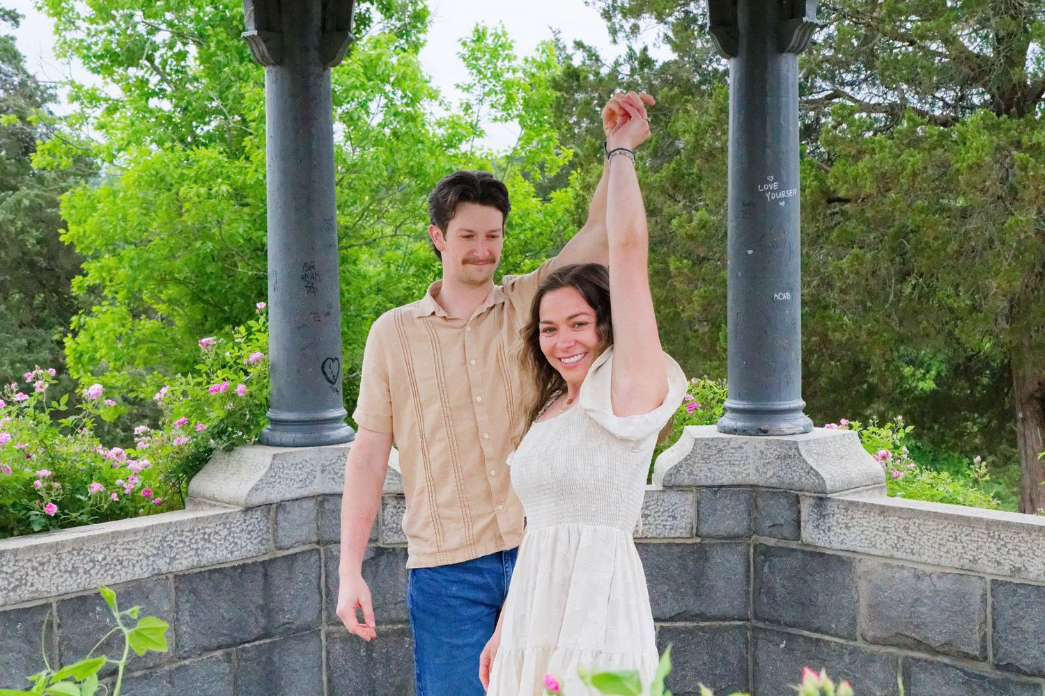 Happy couple posing outdoors under stone columns surrounded by lush green gardens, woman's arm raised joyfully.
