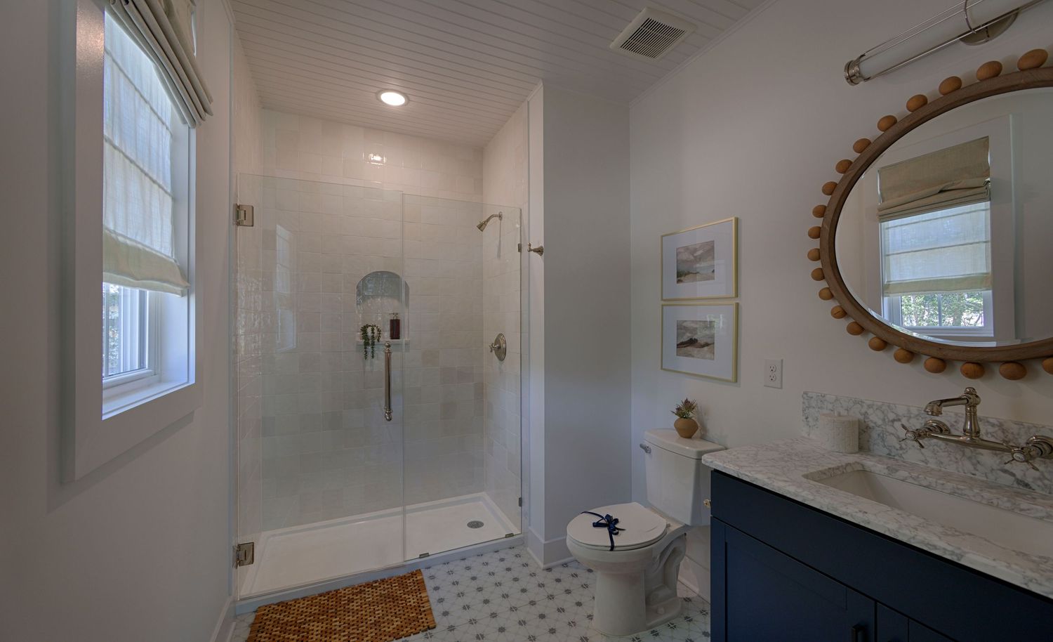 Modern bathroom with glass shower, navy vanity, round mirror, white tile floors, and natural light from windows.