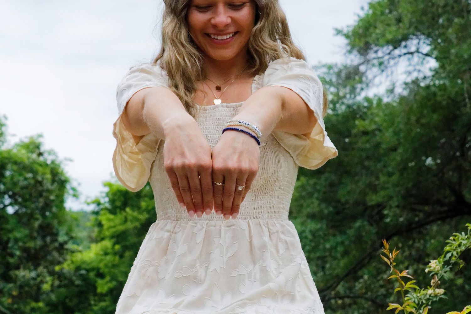 Smiling woman in white dress extending arms forward, showcasing rings and bracelets, outdoors with green trees.