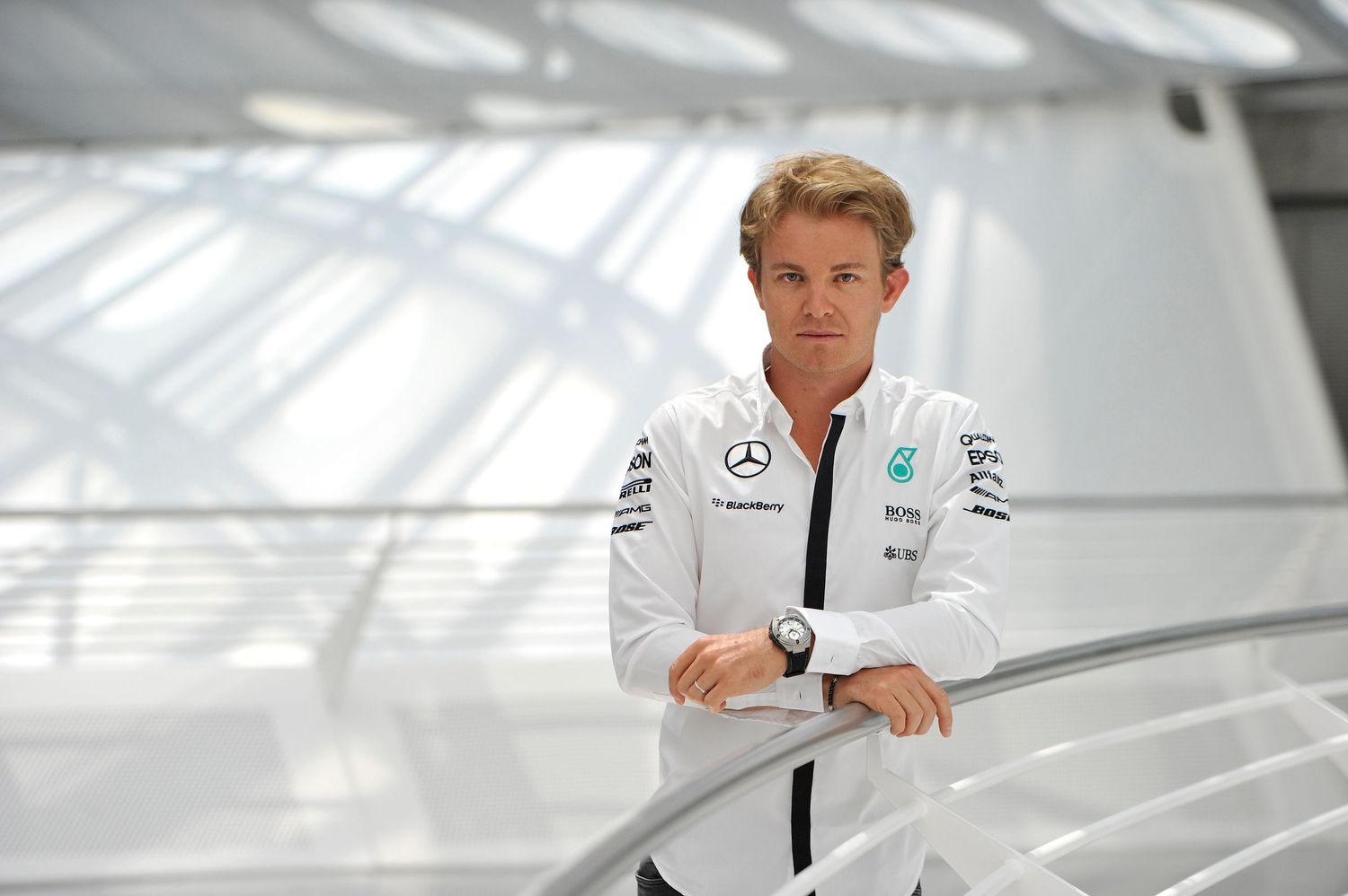 Nico Rosberg portrait in Mercedes-AMG Petronas F1 team white polo shirt leaning on railing indoors.